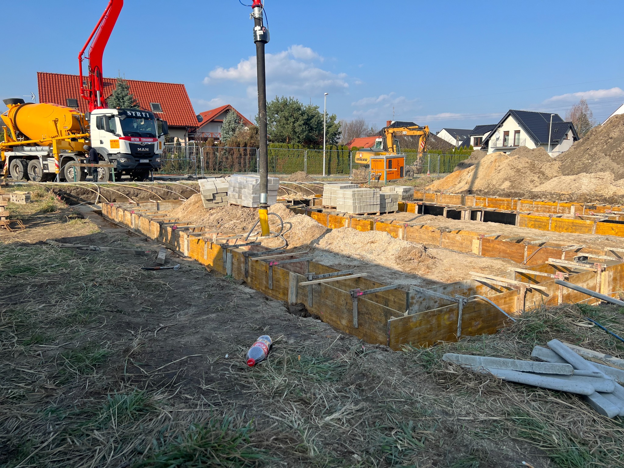 Przygotowanie wykopu pod fundamenty z szalunkami i betonowozem na budowie domu jednorodzinnego w słoneczny dzień; w tle widoczna koparka i zabudowa mieszkalna.