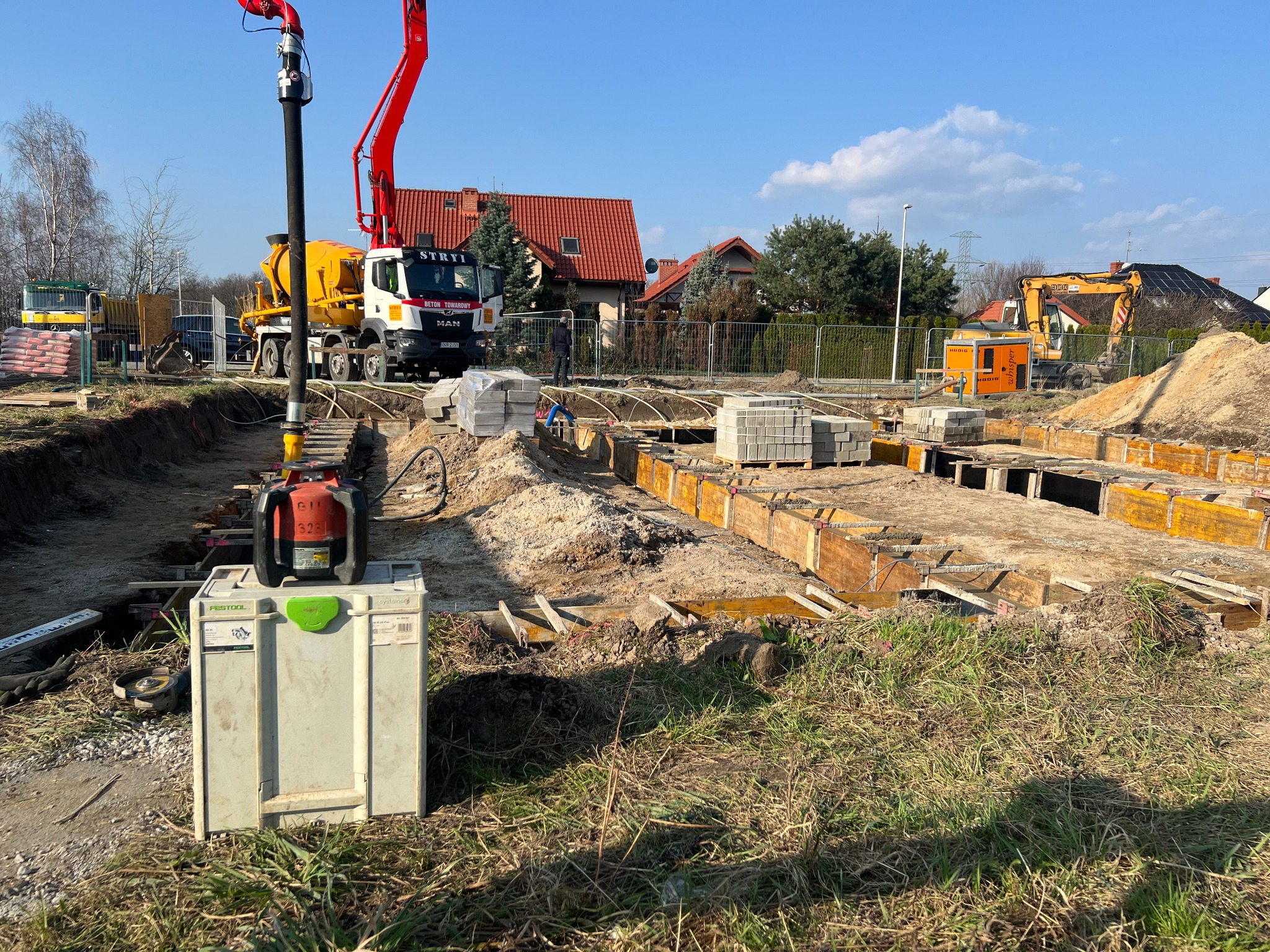 Przygotowanie terenu pod budowę: wykop z drewnianym szalunkiem, pompa do betonu na podwoziu MAN, palety z bloczkami betonowymi i laser niwelacyjny Festool na skrzynce.