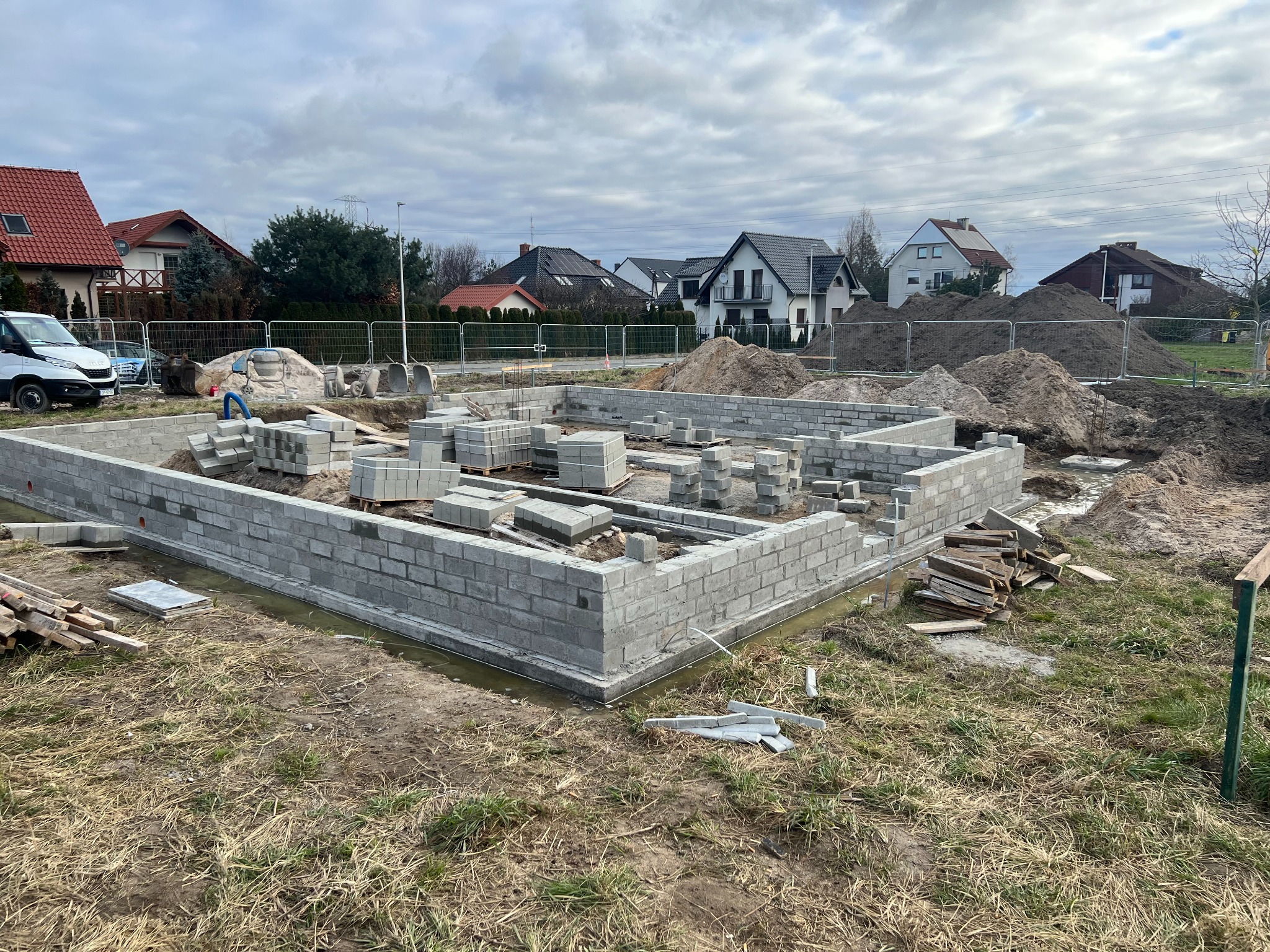 Fundamenty domu z bloczków betonowych w Opolu, częściowo wymurowane ściany, przygotowane bloczki i ziemia w tle, widok na osiedle domów.