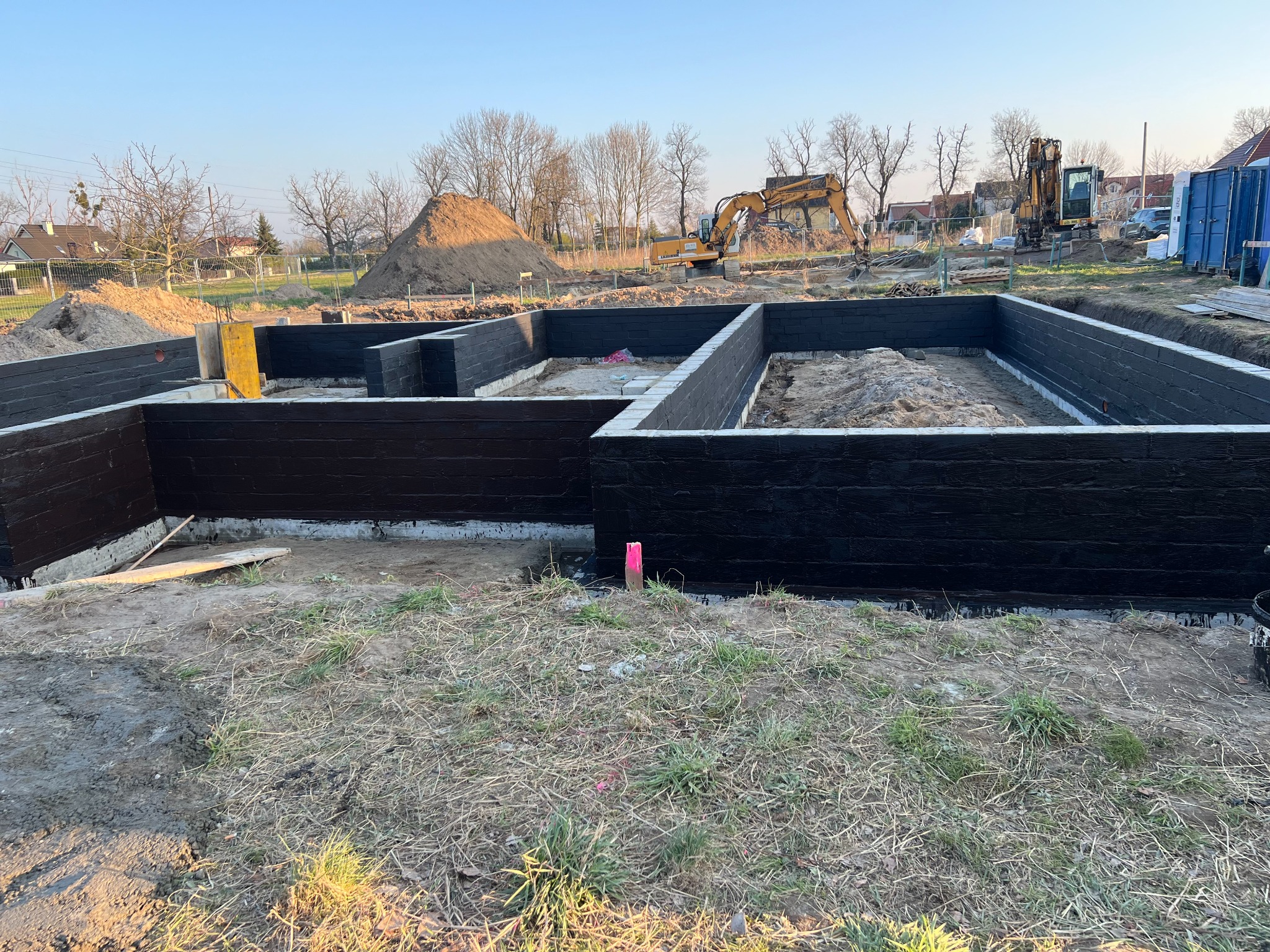 Zabezpieczone fundamenty domu jednorodzinnego, ściany pomalowane na czarno, w tle koparka i hałda ziemi, widok z poziomu gruntu.