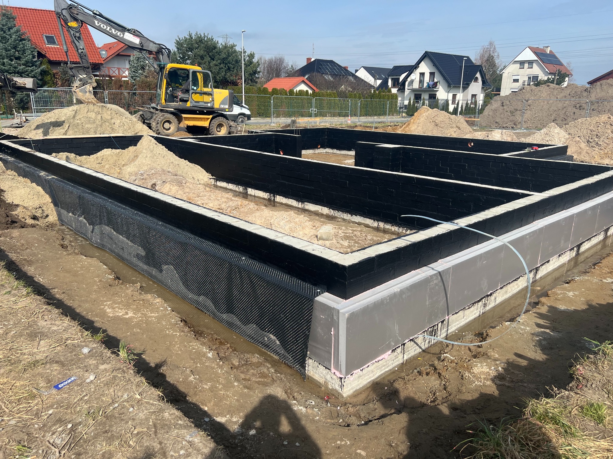 Przygotowany szalunek fundamentów z izolacją pionową i drenażem, na placu budowy stoi żółta koparka Volvo. Widoczne domy jednorodzinne w tle.