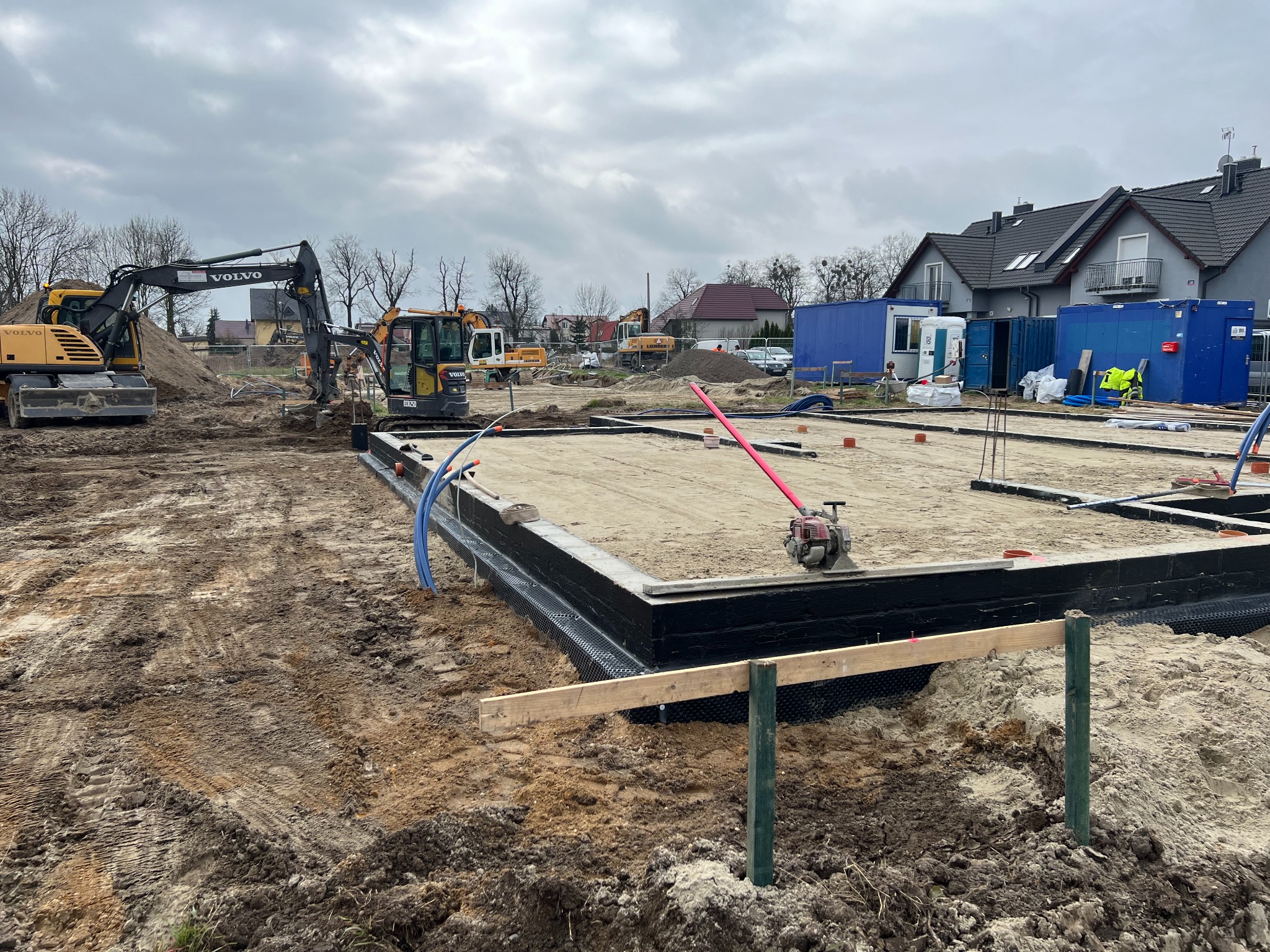 Przygotowany szalunek z izolacją pionową fundamentów pod budowę domu jednorodzinnego, zagęszczarka wibracyjna na piasku, w tle koparki Volvo i kontenery budowlane.
