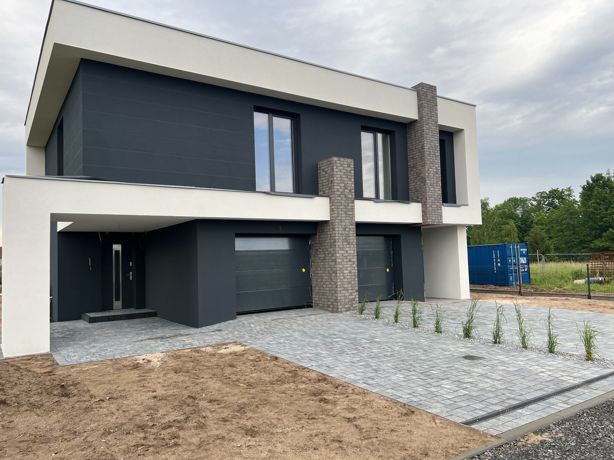Nowoczesny dom murowany z ciemnoszarą elewacją i białymi elementami, kostka brukowa na podjeździe, widoczny kontener budowlany w tle.