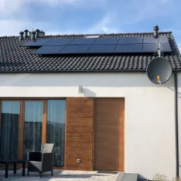 Czarny panel słoneczny na dachu nowoczesnego domu jednorodzinnego z szarą dachówką, widoczna antena satelitarna i wentylatory dachowe, na pierwszym planie taras z meblami ogrodowymi.