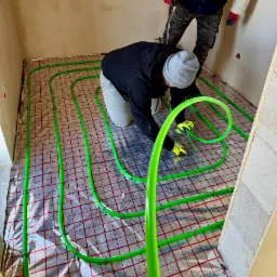 Instalacja ogrzewania podłogowego: instalator w szarej czapce układa zielone rury na folii z czerwoną kratką w surowym wnętrzu, drugi instalator w tle.