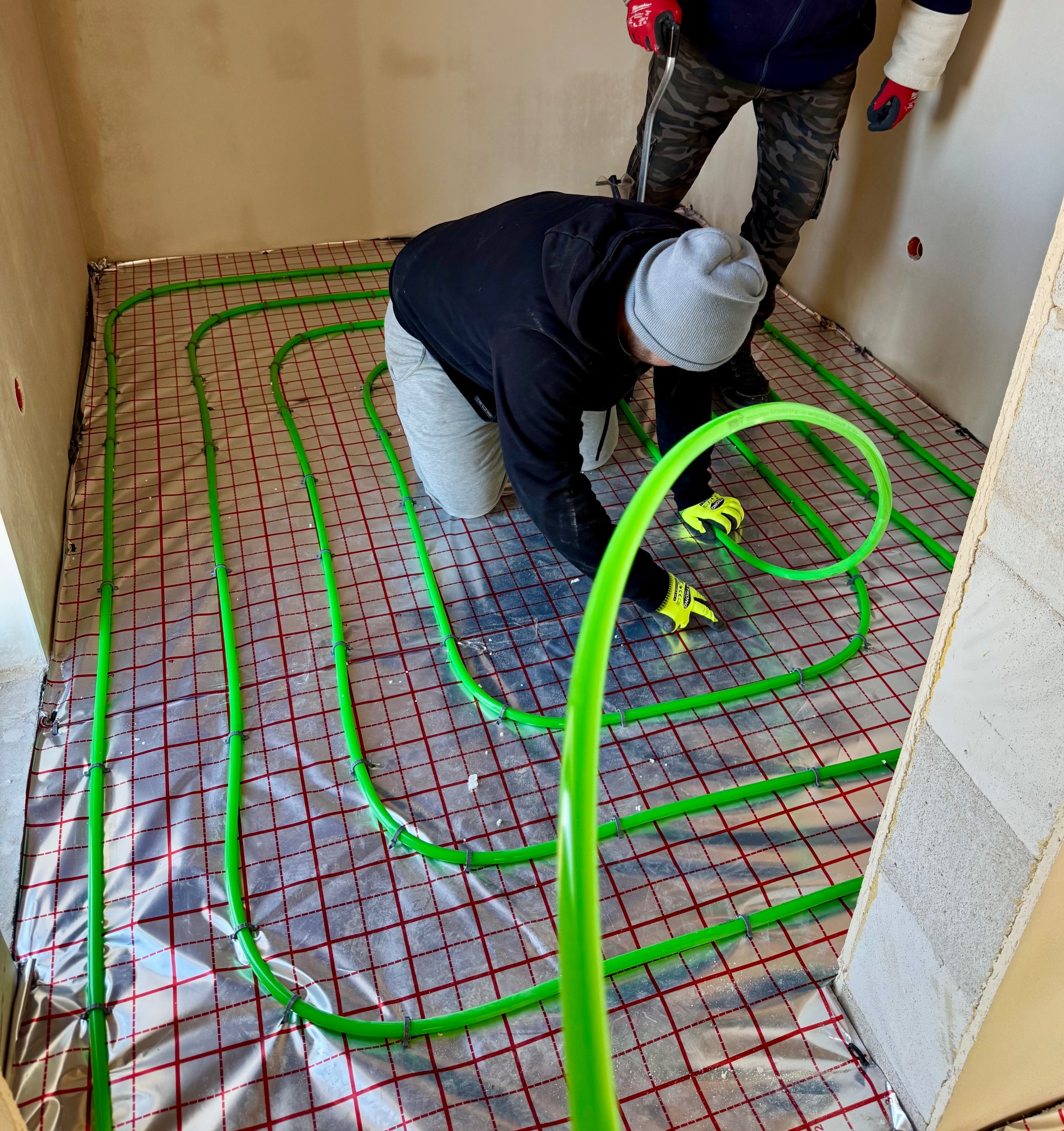 Instalacja ogrzewania podłogowego: instalator w szarej czapce układa zielone rury na folii z czerwoną kratką w surowym wnętrzu, drugi instalator w tle.