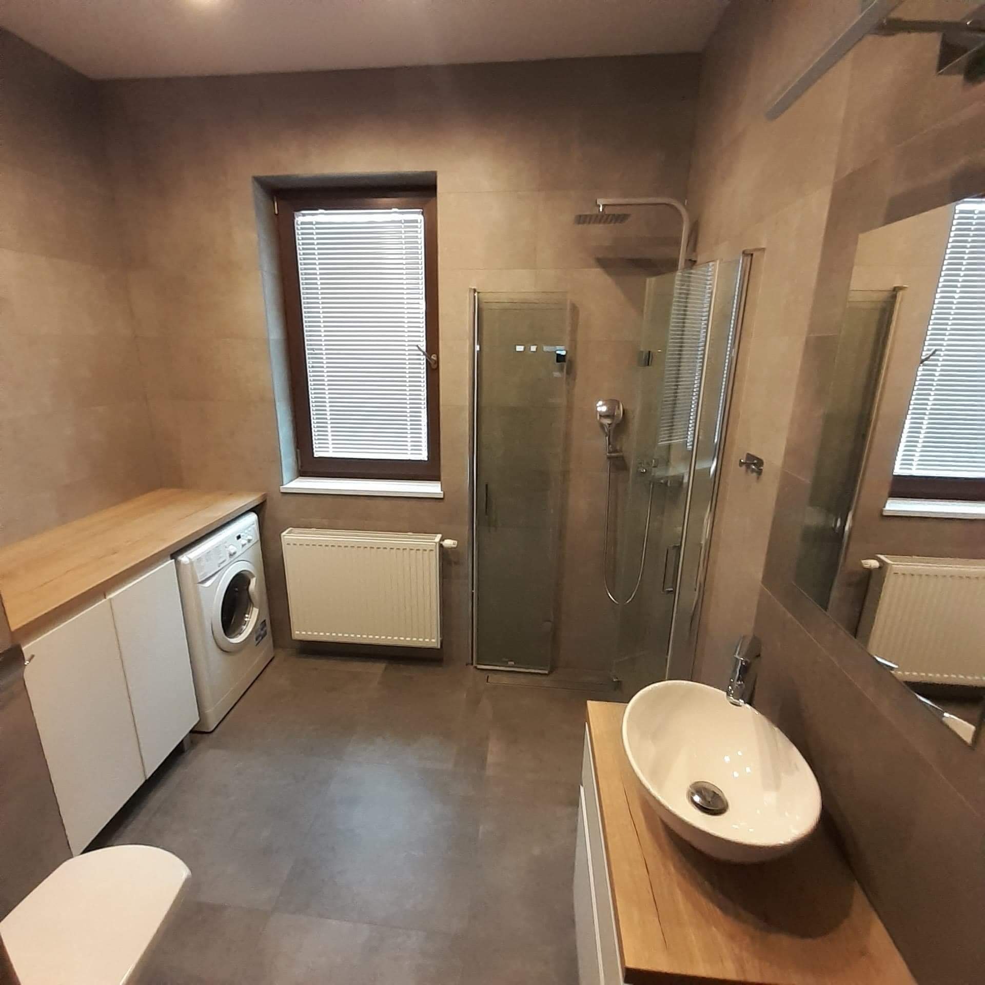 Nowoczesna łazienka w odcieniach beżu z kabiną prysznicową, umywalką nablatową na drewnianym blacie, pralką zabudowaną w szafce i grzejnikiem pod oknem z żaluzjami.