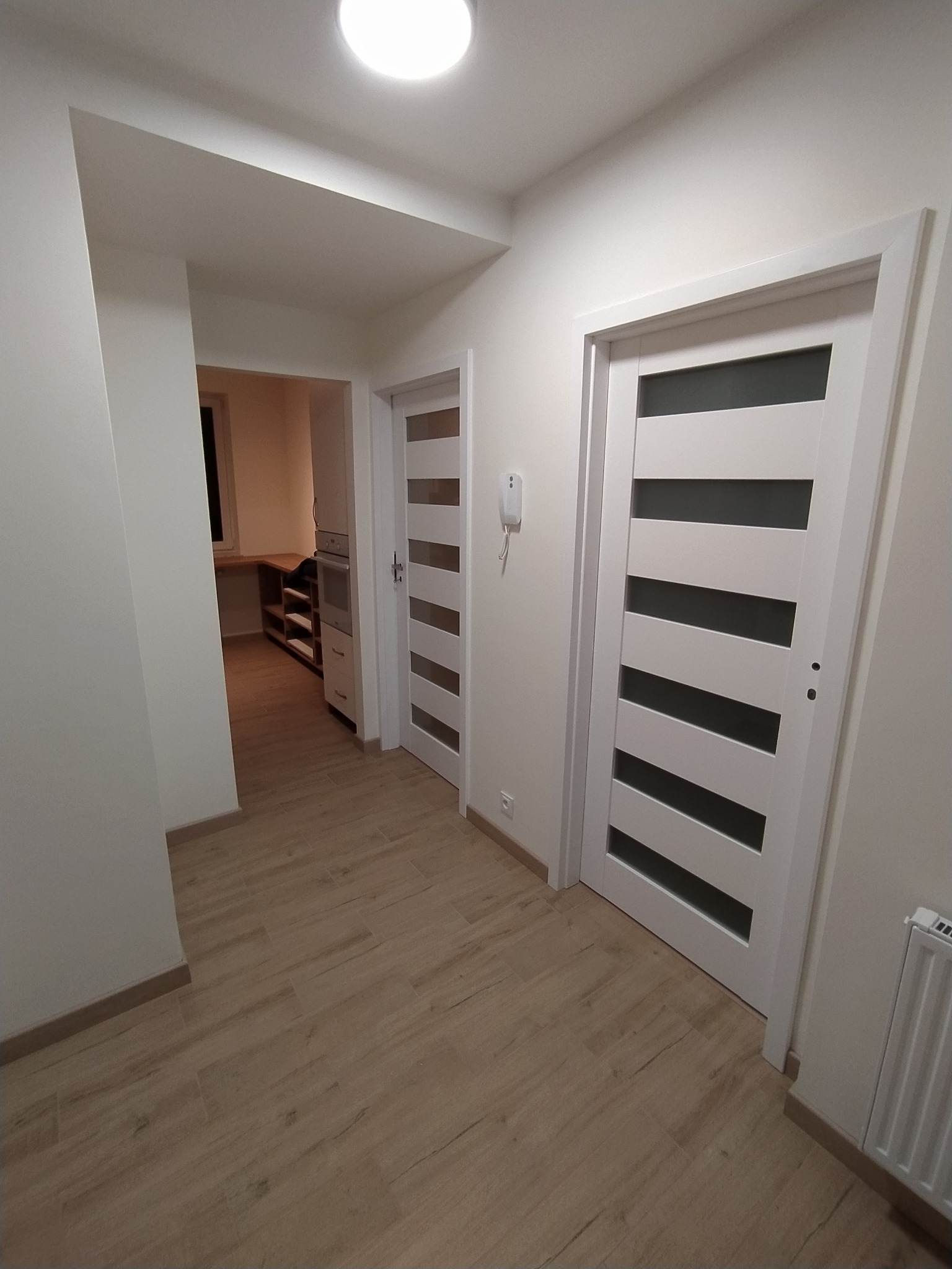 Korytarz z dwiema białymi drzwiami z poziomymi przeszkleniami, drewnopodobna podłoga, białe ściany, widoczny fragment grzejnika i interkom. W tle widoczne wejście do kuchni z zabudową meblową.