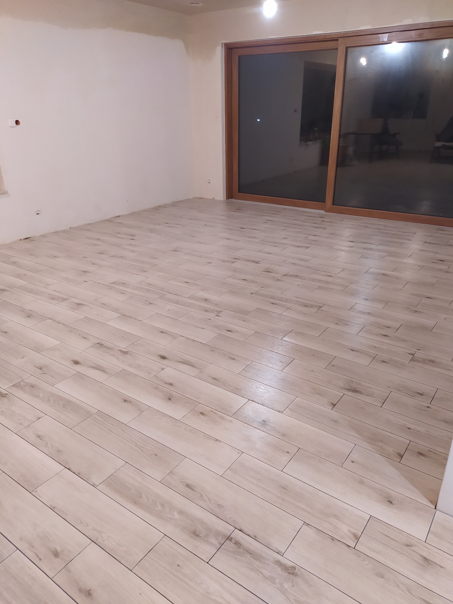 Jasne, podłużne panele ułożone w nowym pokoju, widoczne przesuwne drzwi balkonowe z brązowymi ramami i przygotowana do wykończenia ściana.