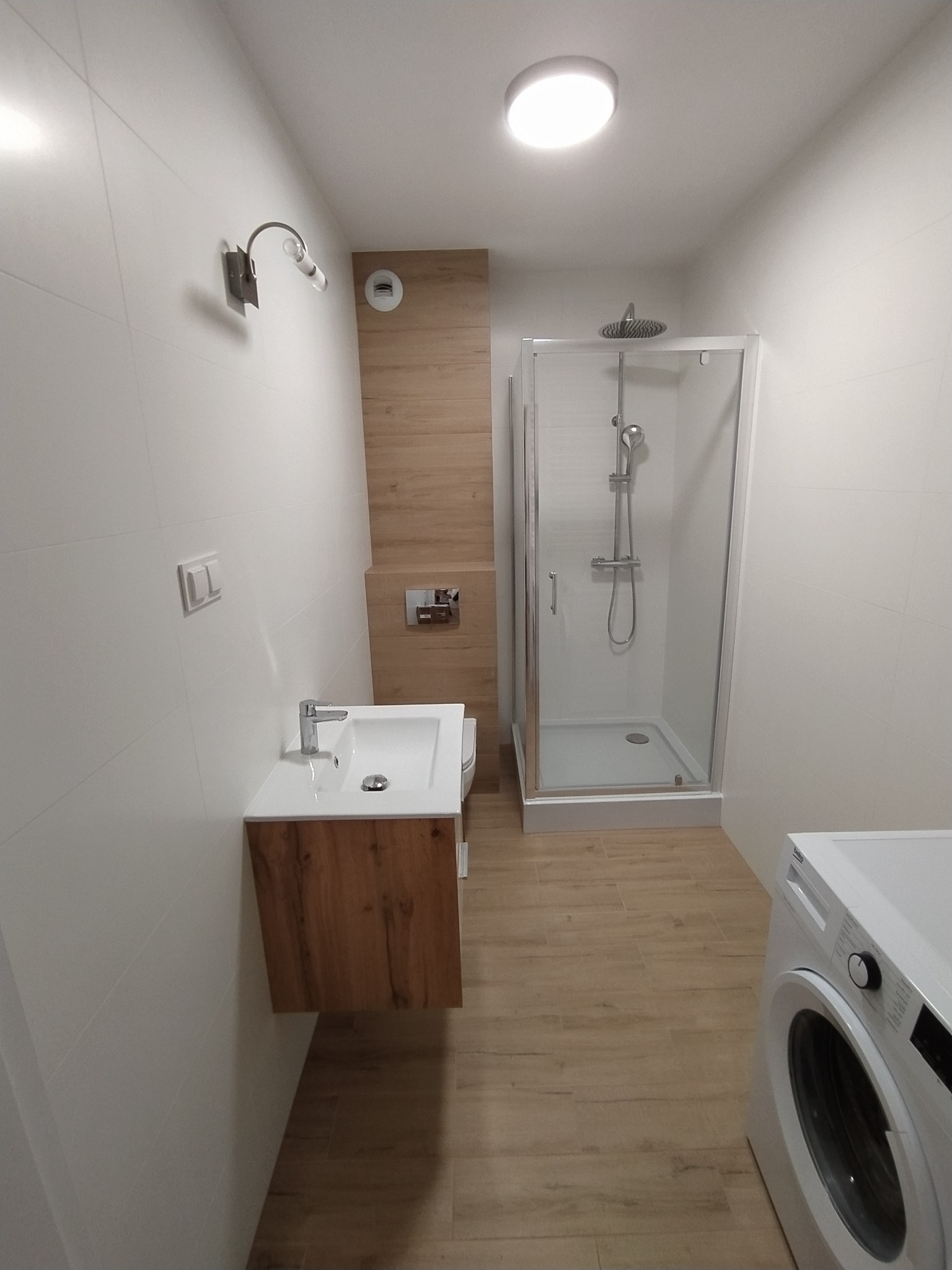 Nowoczesna łazienka po remoncie: białe płytki, drewnopodobna podłoga, kabina prysznicowa ze srebrnym wykończeniem, umywalka z szafką w kolorze drewna, pralka.