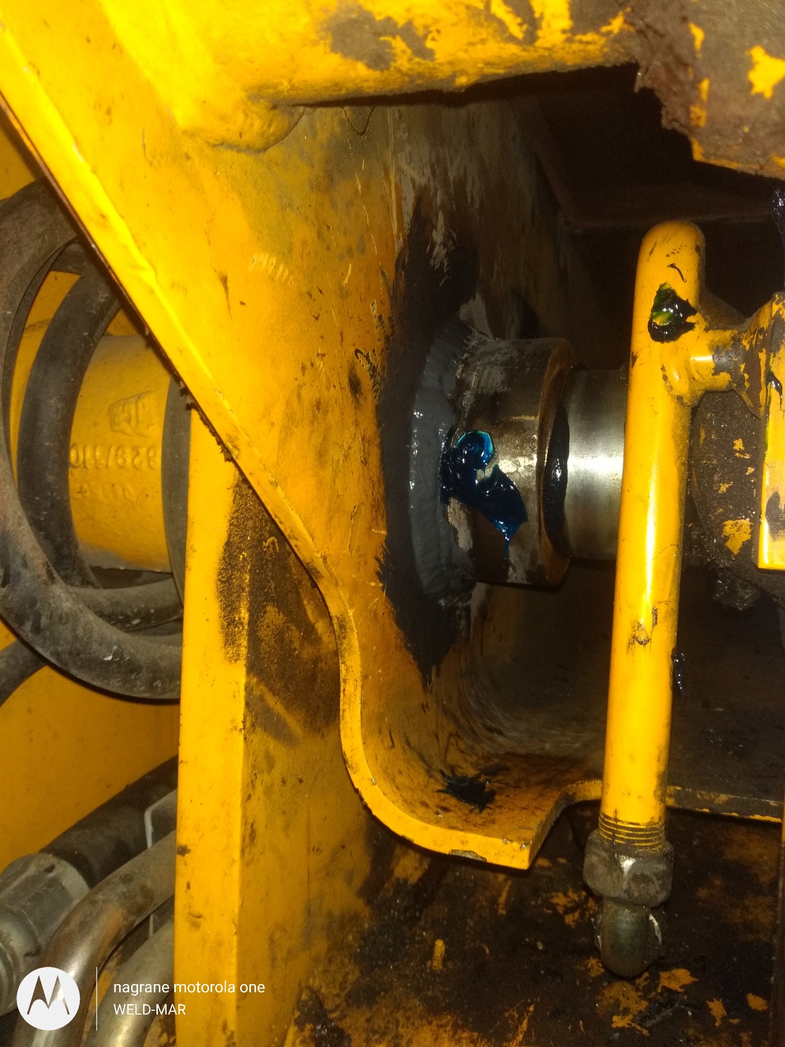Detal widoku smarowania łożyska w żółtej konstrukcji stalowej maszyny budowlanej, z widocznymi śladami użytkowania i ciemnym smarem.