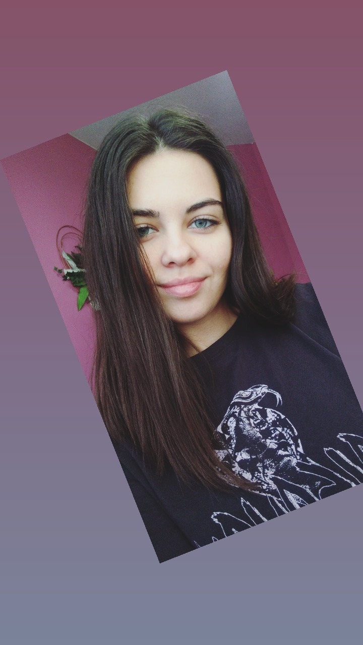 Portret uśmiechniętej kobiety z heterochromią, ciemnymi włosami i czarnym T-shirtem z nadrukiem, na tle różowej ściany z dekoracją roślinną.