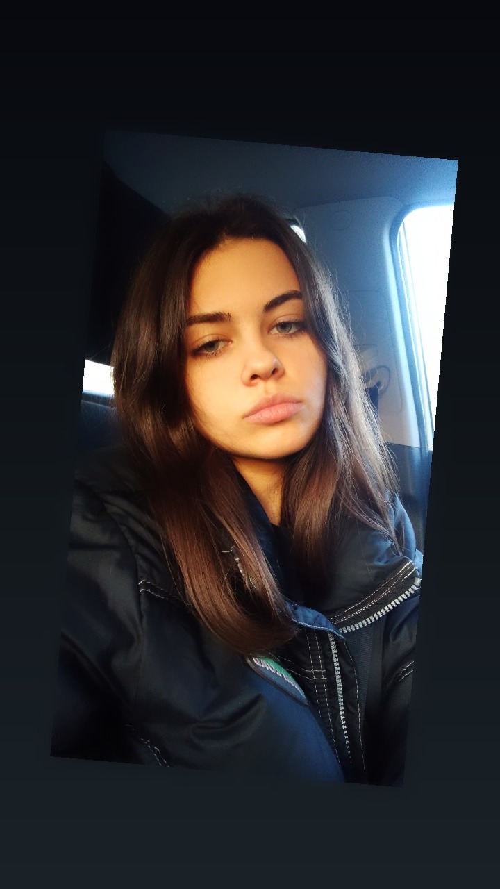 Portret młodej kobiety z brązowymi włosami i niebieskimi oczami w ciemnej kurtce, siedzącej w samochodzie.