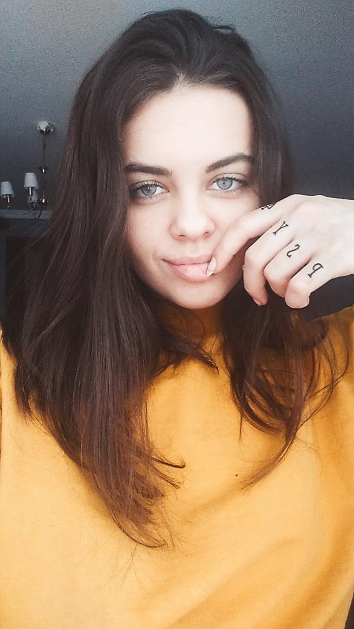 Portret młodej kobiety o jasnych oczach i ciemnych włosach, ubrana w żółty golf, z tatuażami na palcach.