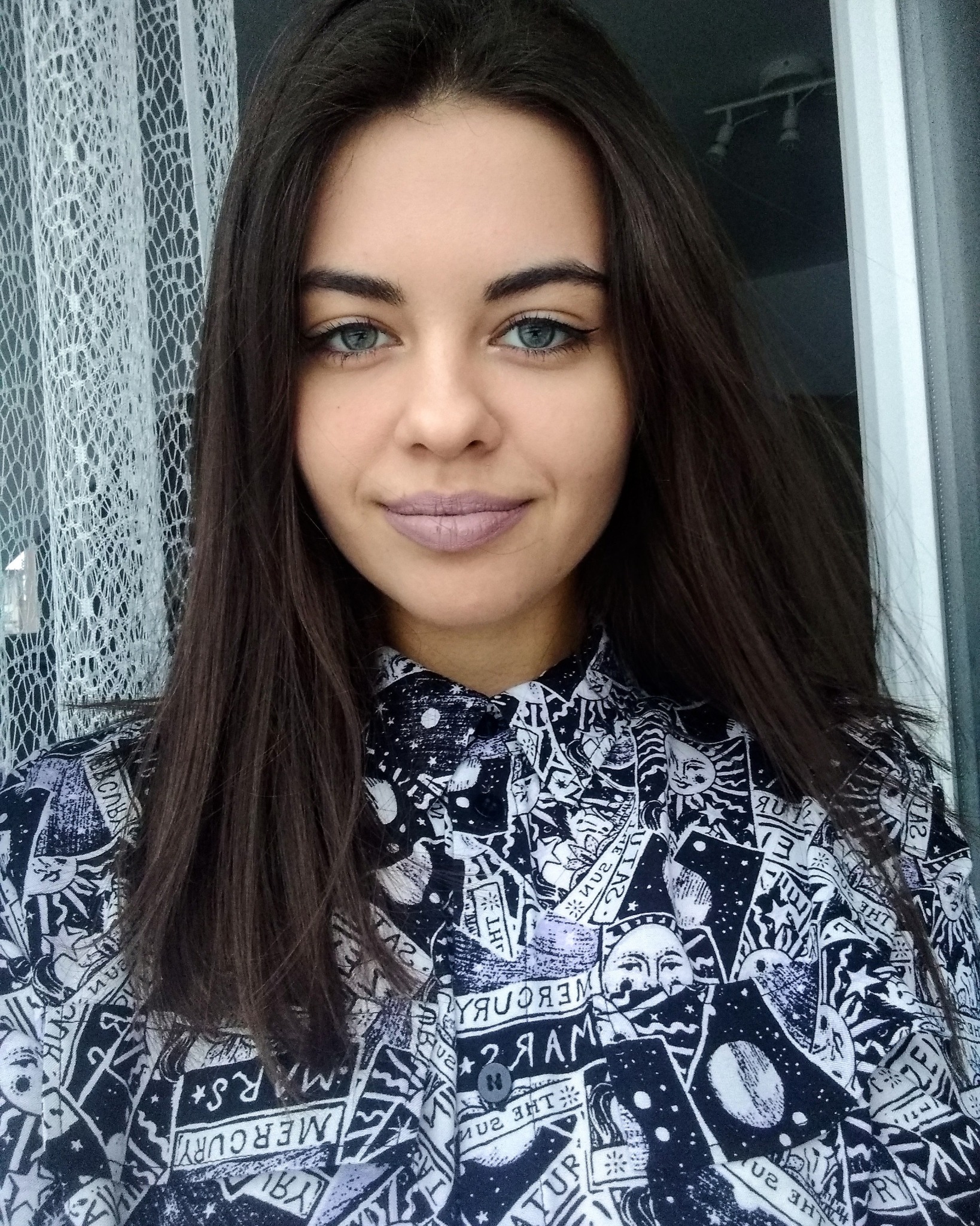 Portret młodej kobiety z długimi ciemnymi włosami, błękitnymi oczami i fioletową szminką, ubrana w koszulę z motywem astrologicznym na tle firanki.