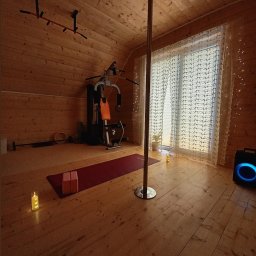 Trener Personalny - Domowa siłownia na poddaszu z drążkiem, atlasem, matą do ćwiczeń i rurą do pole dance. Drewniane wykończenie, okno z firankami i nastrojowe oświetlenie.