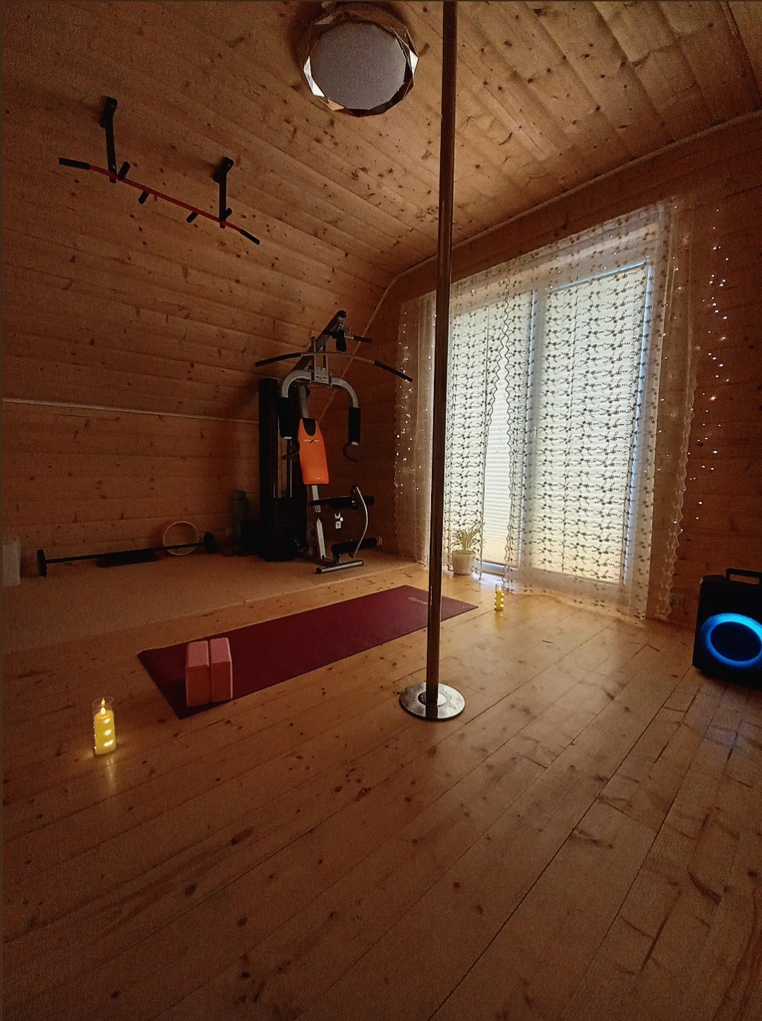 Domowa siłownia na poddaszu z drążkiem, atlasem, matą do ćwiczeń i rurą do pole dance. Drewniane wykończenie, okno z firankami i nastrojowe oświetlenie.