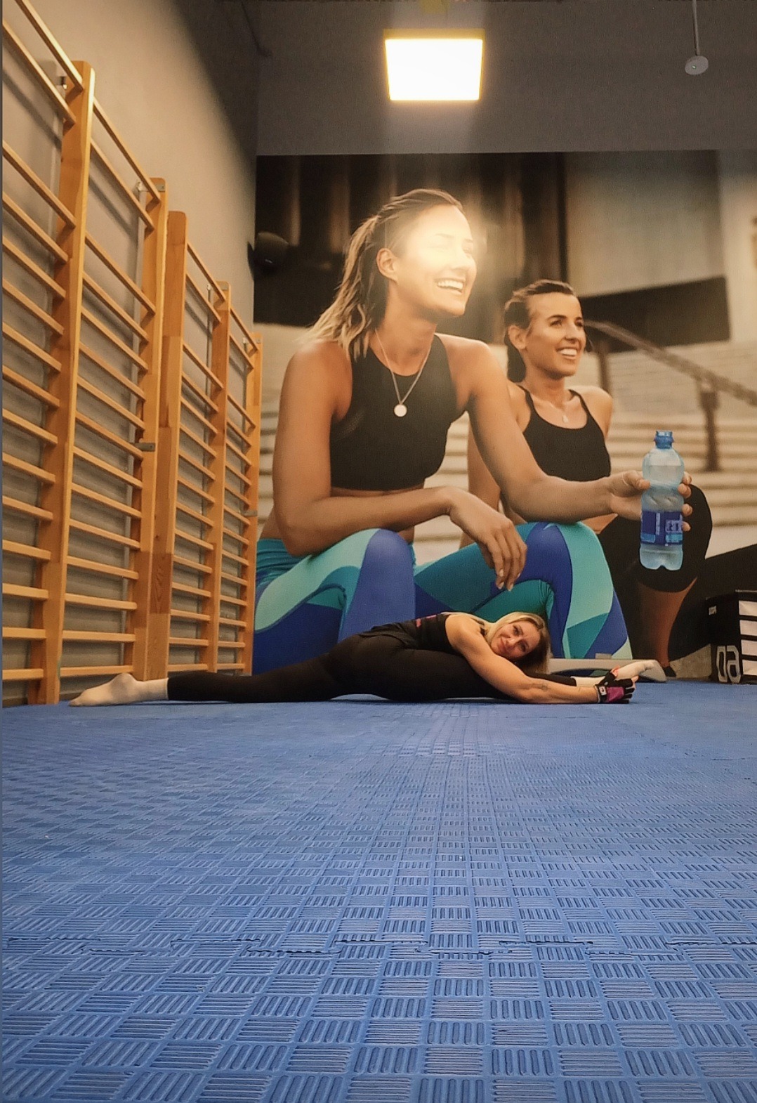 Kobieta w rozkroku na niebieskiej macie, w tle drabinki gimnastyczne i plakat z uśmiechniętymi kobietami w strojach sportowych. Trening rozciągający w jasnym pomieszczeniu.