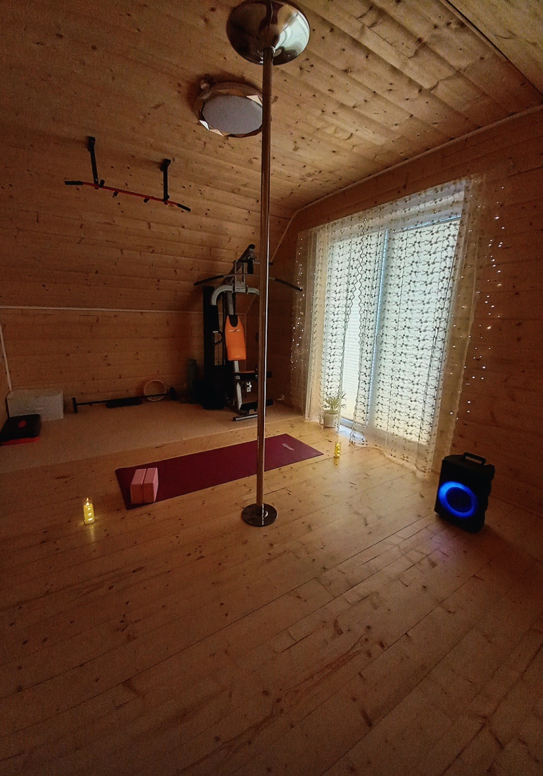 Domowa siłownia na poddaszu z drążkiem do pole dance, matą do ćwiczeń i sprzętem fitness. Drewniane ściany i podłoga tworzą przytulną atmosferę.