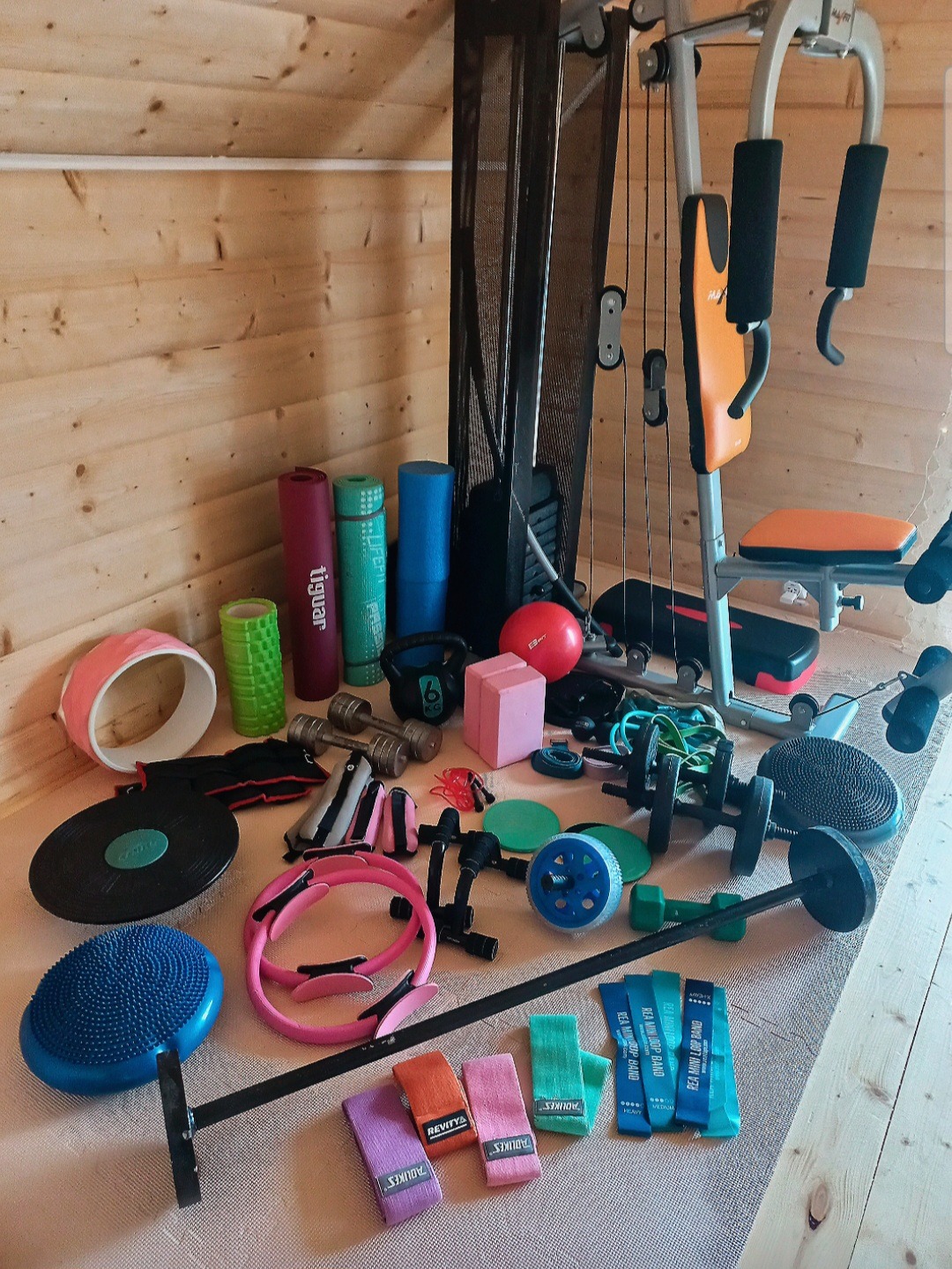 Różnorodny sprzęt fitness: hantle, maty, gumy oporowe, piłki i maszyna do ćwiczeń na tle drewnianej ściany. Bogaty zestaw akcesoriów do treningu personalnego.