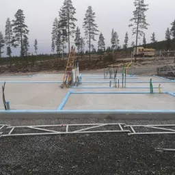 Betonowa płyta fundamentowa z niebieską izolacją obwodową, wystającymi rurami instalacyjnymi i drabiną opartą o skrzynkę elektryczną na tle lasu i pochmurnego nieba.