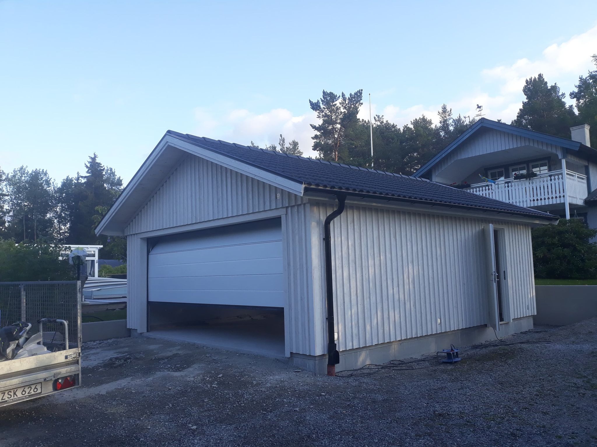 Nowoczesny garaż z częściowo otwartą segmentową bramą wjazdową, obok przyczepa z narzędziami i materiałami budowlanymi.