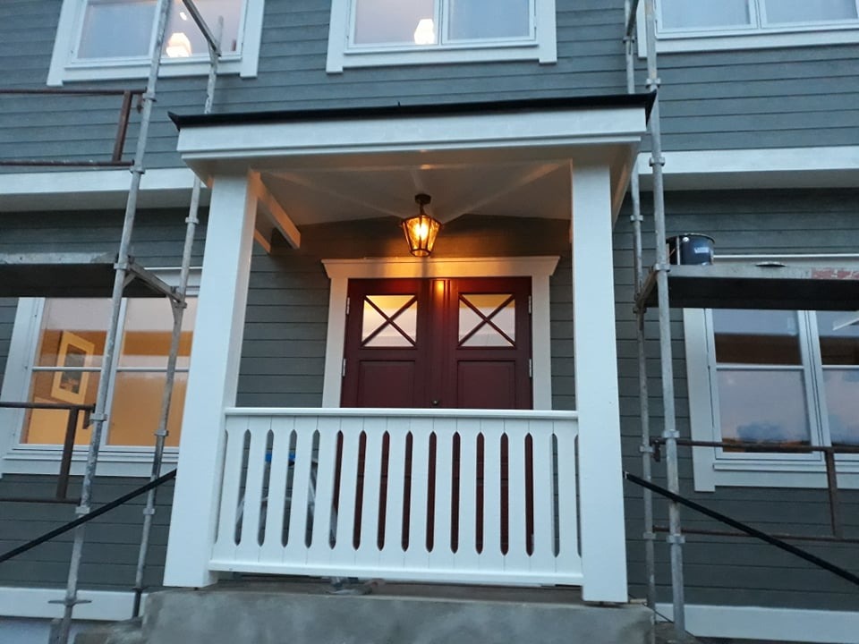 Wejście do domu z drewnianą werandą w trakcie prac, widoczne rusztowania i wiadro z farbą, szare deski elewacyjne.