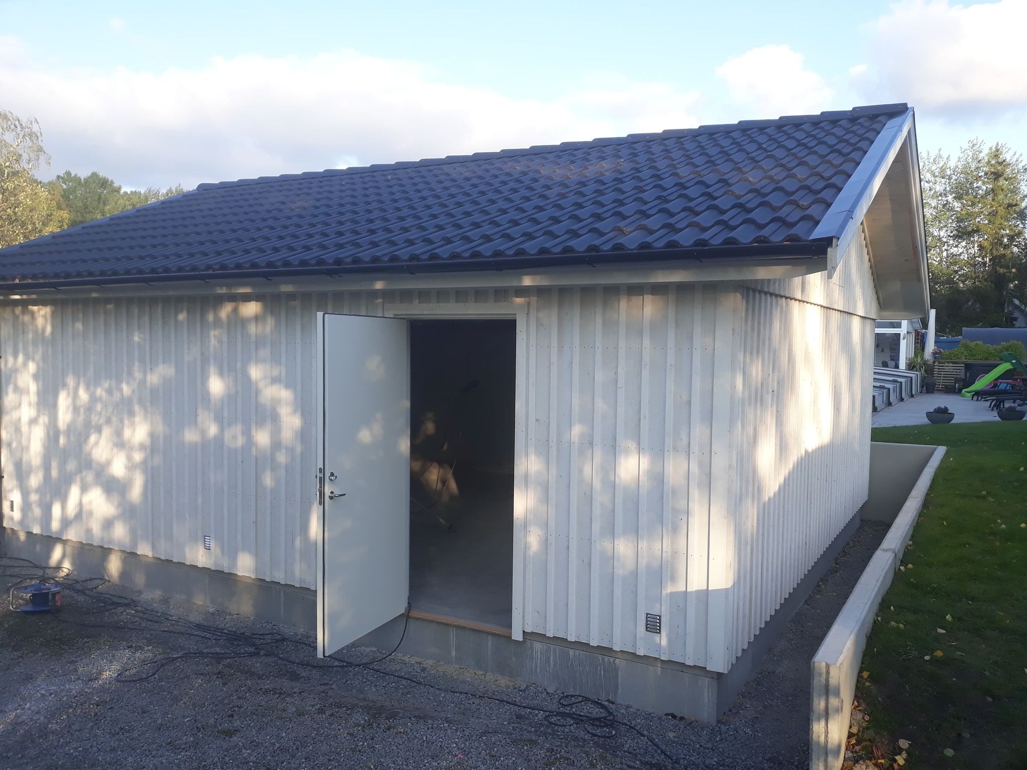 Nowo wybudowany, biały budynek gospodarczy z czarnym dachem z blachodachówki, otwartymi drzwiami i widocznymi przewodami elektrycznymi na żwirowym podłożu.