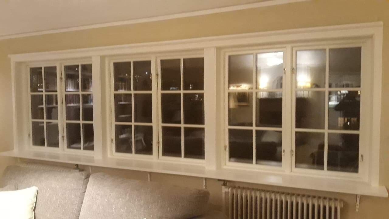 Trzy białe, wielokwaterowe okna z parapetem wewnątrz pomieszczenia, widoczny fragment sofy i grzejnika, nocne odbicie wnętrza w szybach.