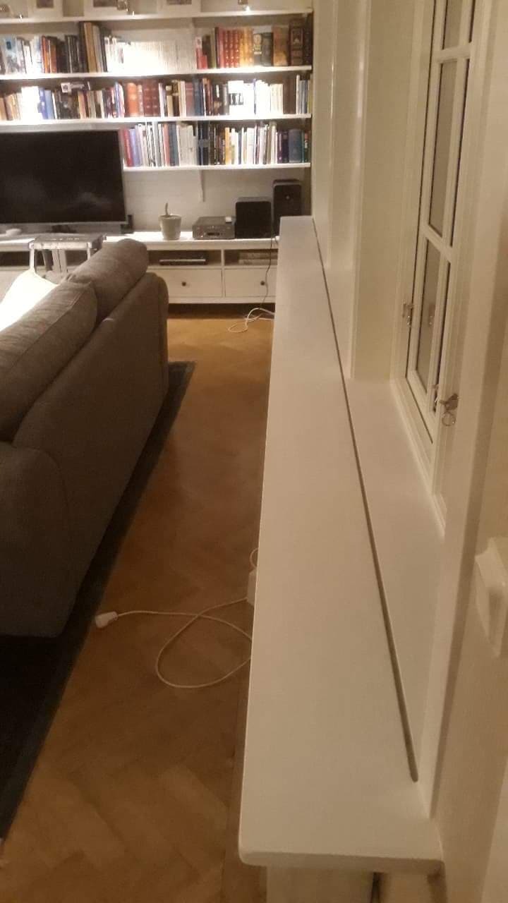 Jasne wnętrze: świeżo pomalowane ściany, okno z białymi ramami i parapetem, parkiet na podłodze, szara sofa w tle, regał z książkami i telewizorem.