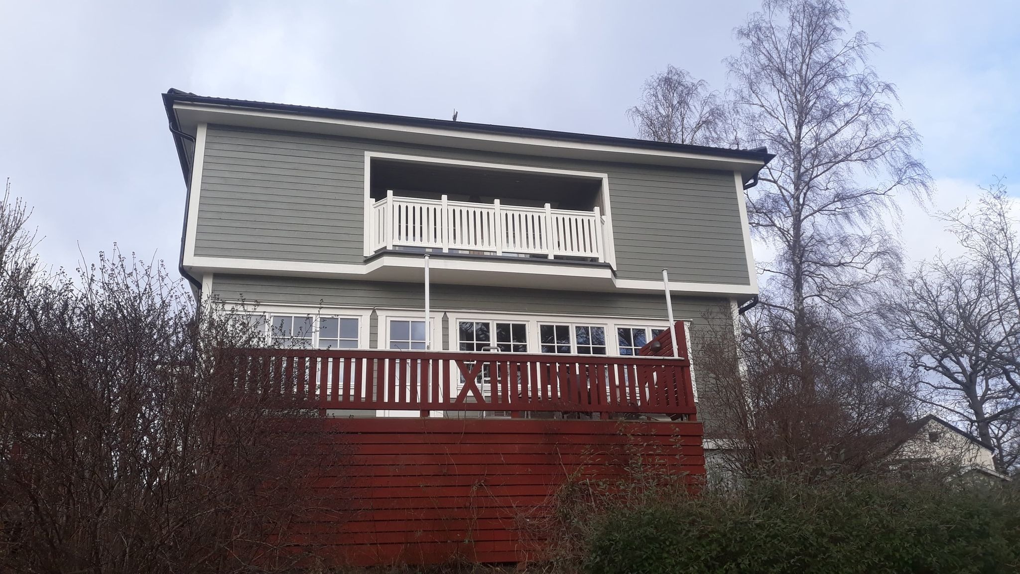 Dwukondygnacyjny dom z szarą elewacją z desek, białymi obramowaniami okien i balkonem z białymi balustradami, z czerwonym tarasem na parterze, widziany zza krzewów.