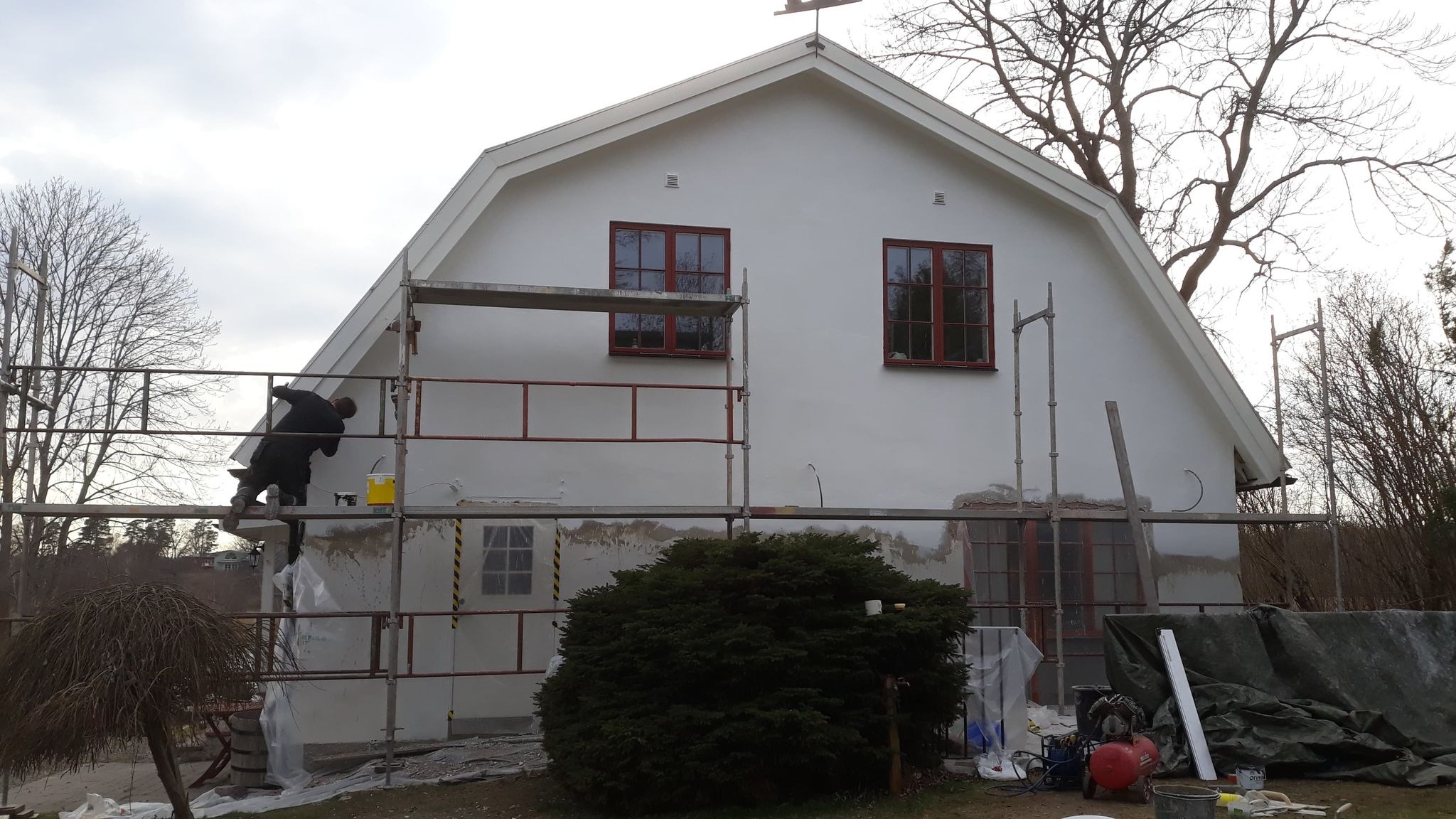 Pracownik na rusztowaniu, maluje elewację dwupiętrowego domu z czerwonymi ramami okiennymi, widoczne ślady wcześniejszych prac i przygotowania do malowania, w tle drzewa bez liści.