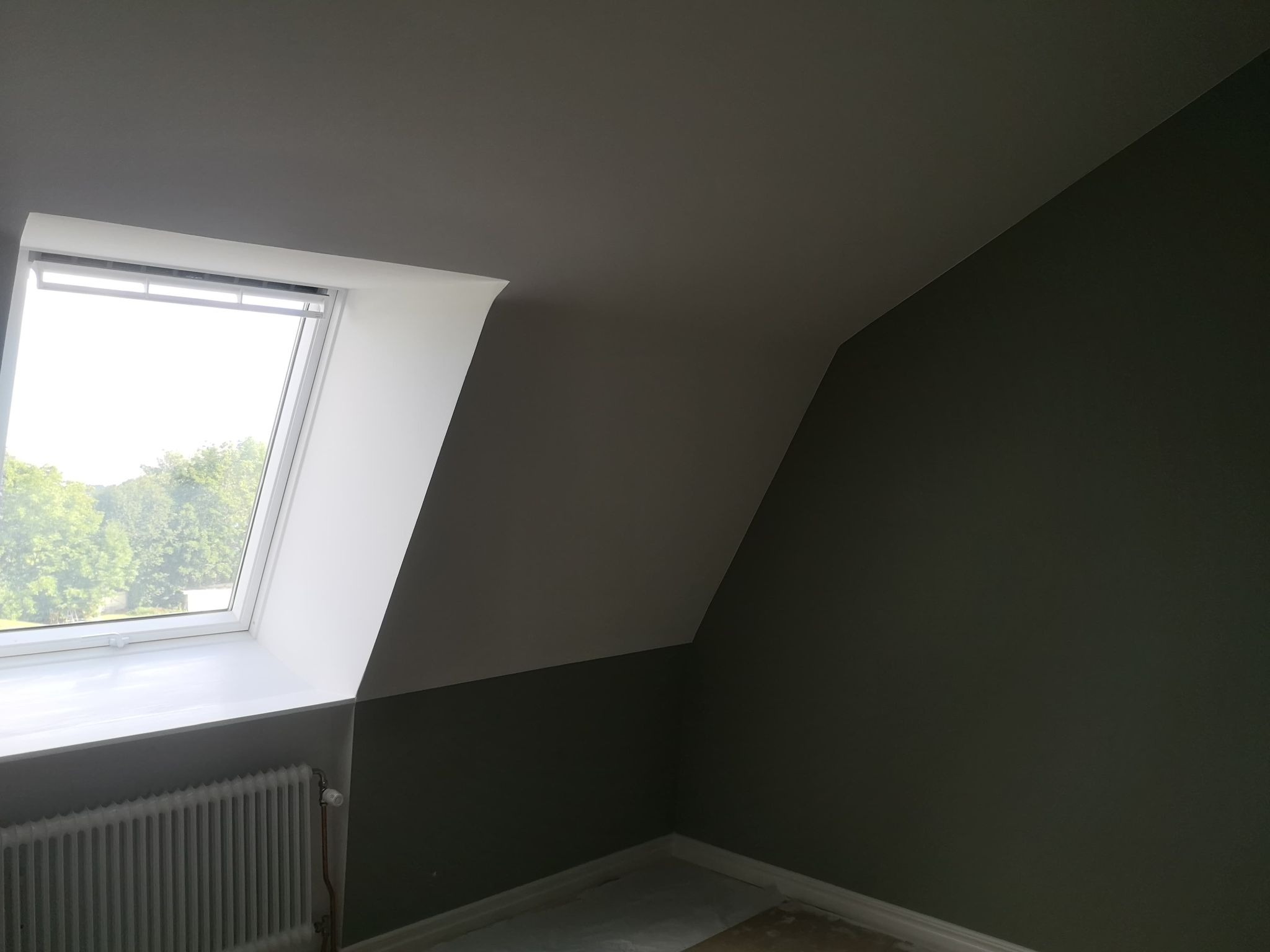 Pomalowane na szaro poddasze z oknem dachowym i grzejnikiem, widok z dołu na skośną ścianę i sufit.