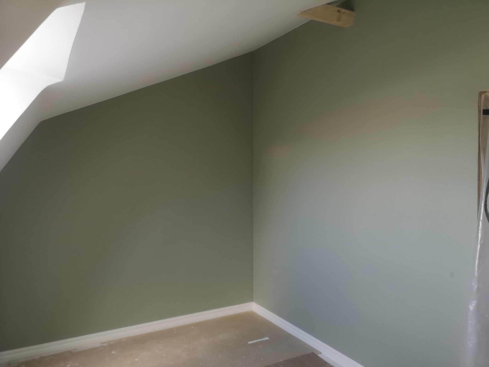Pomalowane na oliwkowy kolor wnętrze poddasza ze skosami, widoczne listwy przypodłogowe i fragment okna dachowego.