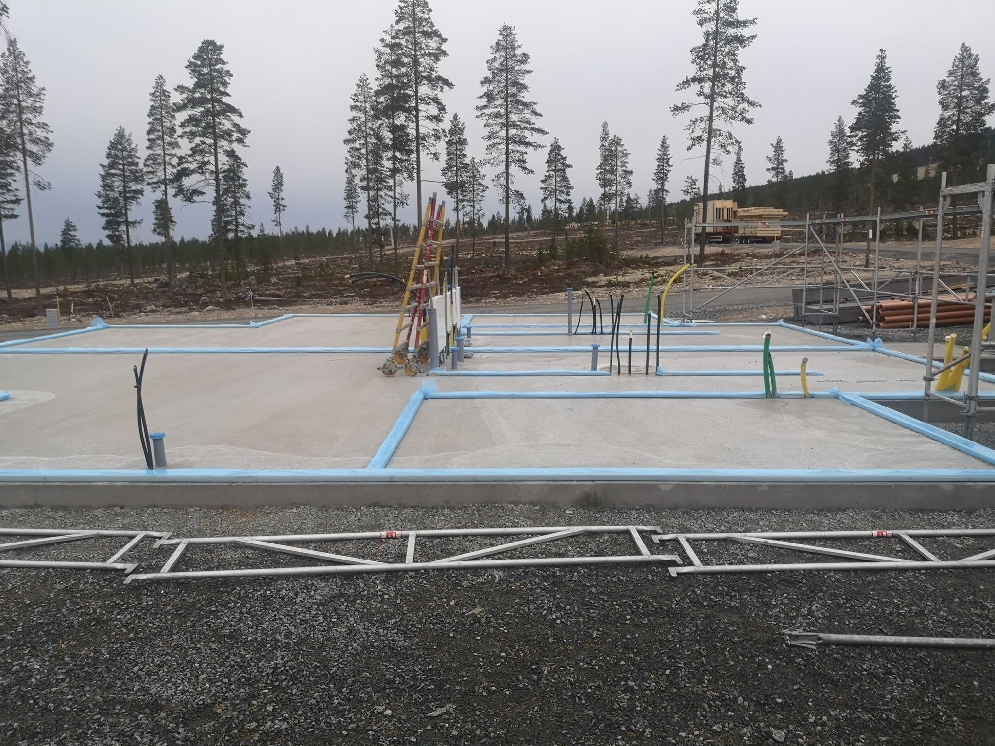 Betonowa płyta fundamentowa z niebieską izolacją obwodową, wystającymi rurami instalacyjnymi i drabiną opartą o skrzynkę elektryczną na tle lasu i pochmurnego nieba.