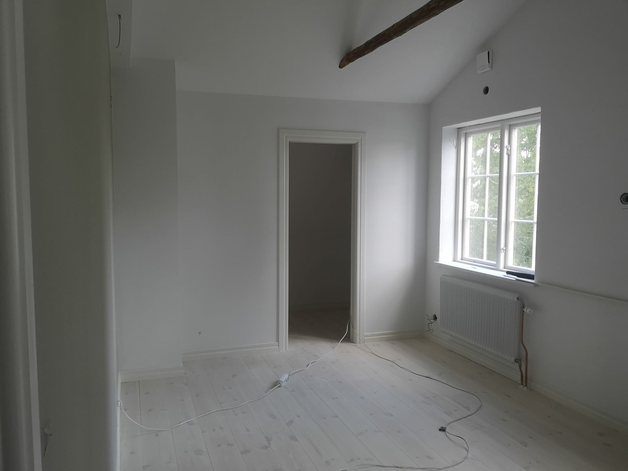 Wnętrze pokoju po odświeżeniu, z białymi ścianami, podłogą i grzejnikiem, widoczne kable elektryczne na podłodze i fragment drewnianej belki na suficie.