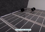 Szare panele grzewcze podłogowe z widocznymi czarnymi elementami instalacji doprowadzającej, ułożone na jasnej podłodze, widok z góry pod kątem.