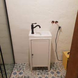 Dim proekt - Mała biała szafka pod umywalkę z czarną baterią na tle białej ściany z wystającymi kablami i wzorzystej podłogi z płytek.