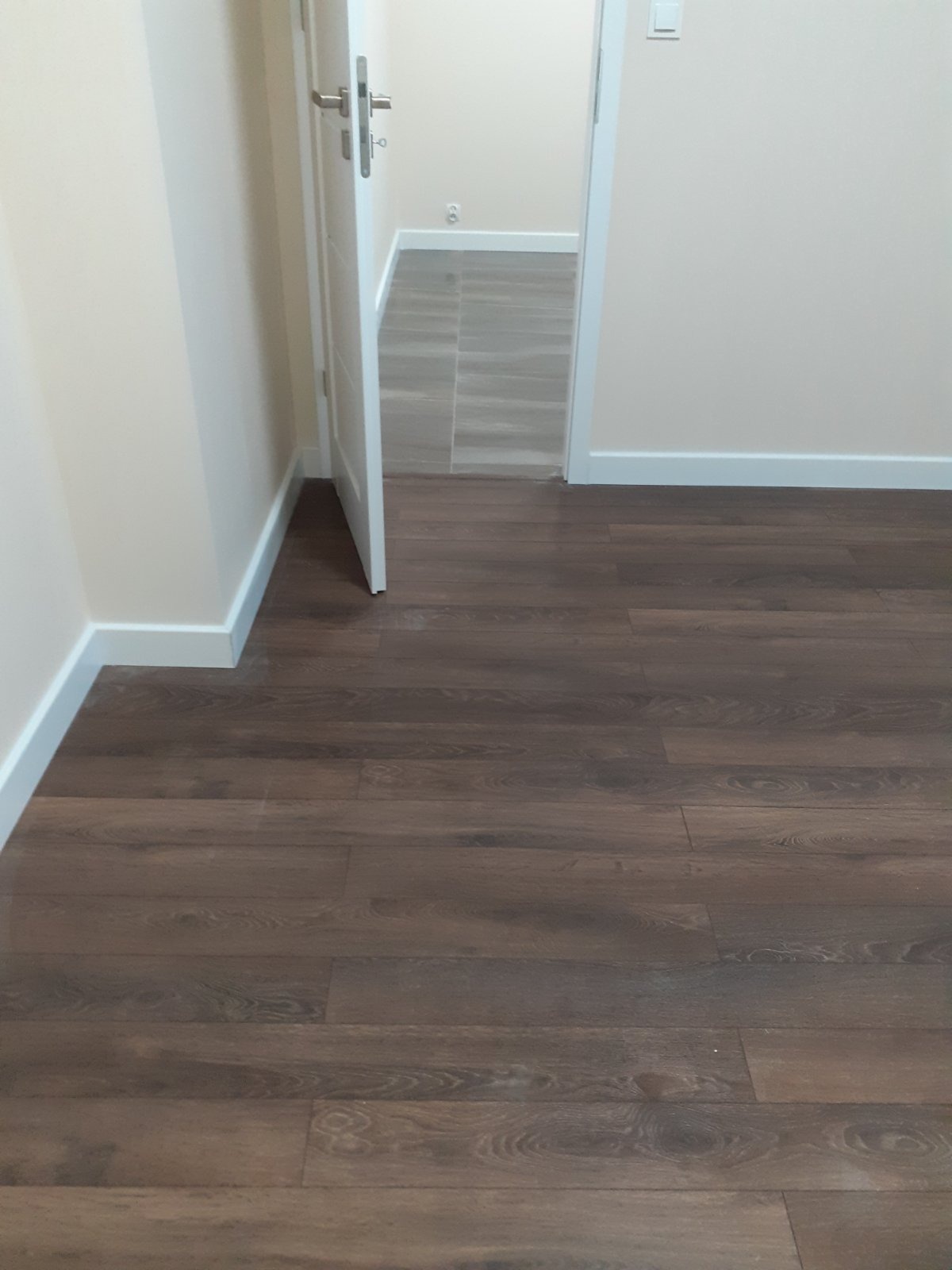 Ułożone ciemne panele podłogowe z widoczną strukturą drewna, białe listwy przypodłogowe i otwarte białe drzwi do kolejnego pomieszczenia z szarą podłogą.