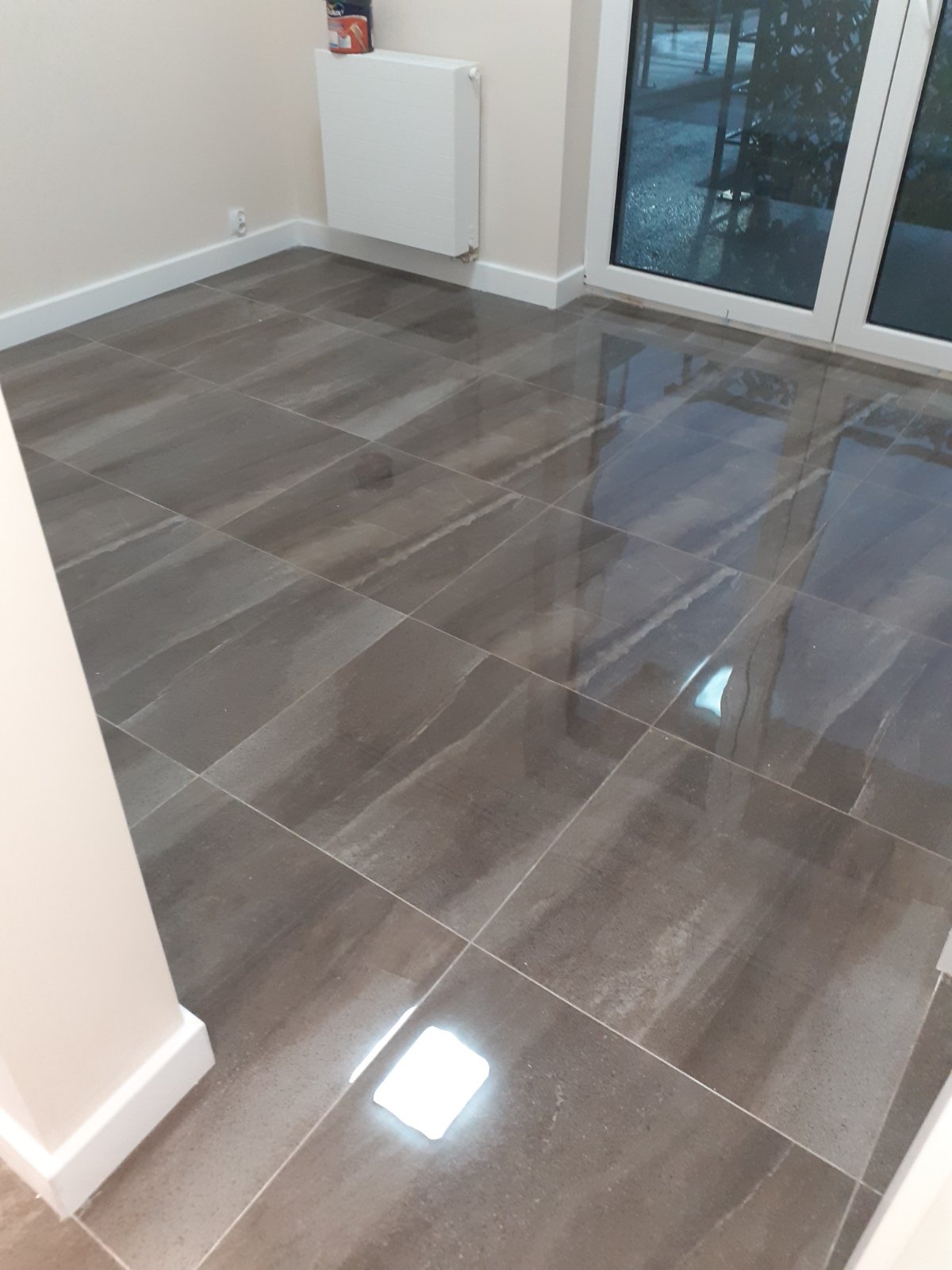 Ułożone ciemnoszare płytki podłogowe z połyskiem, widoczne białe listwy przypodłogowe i fragment okna.