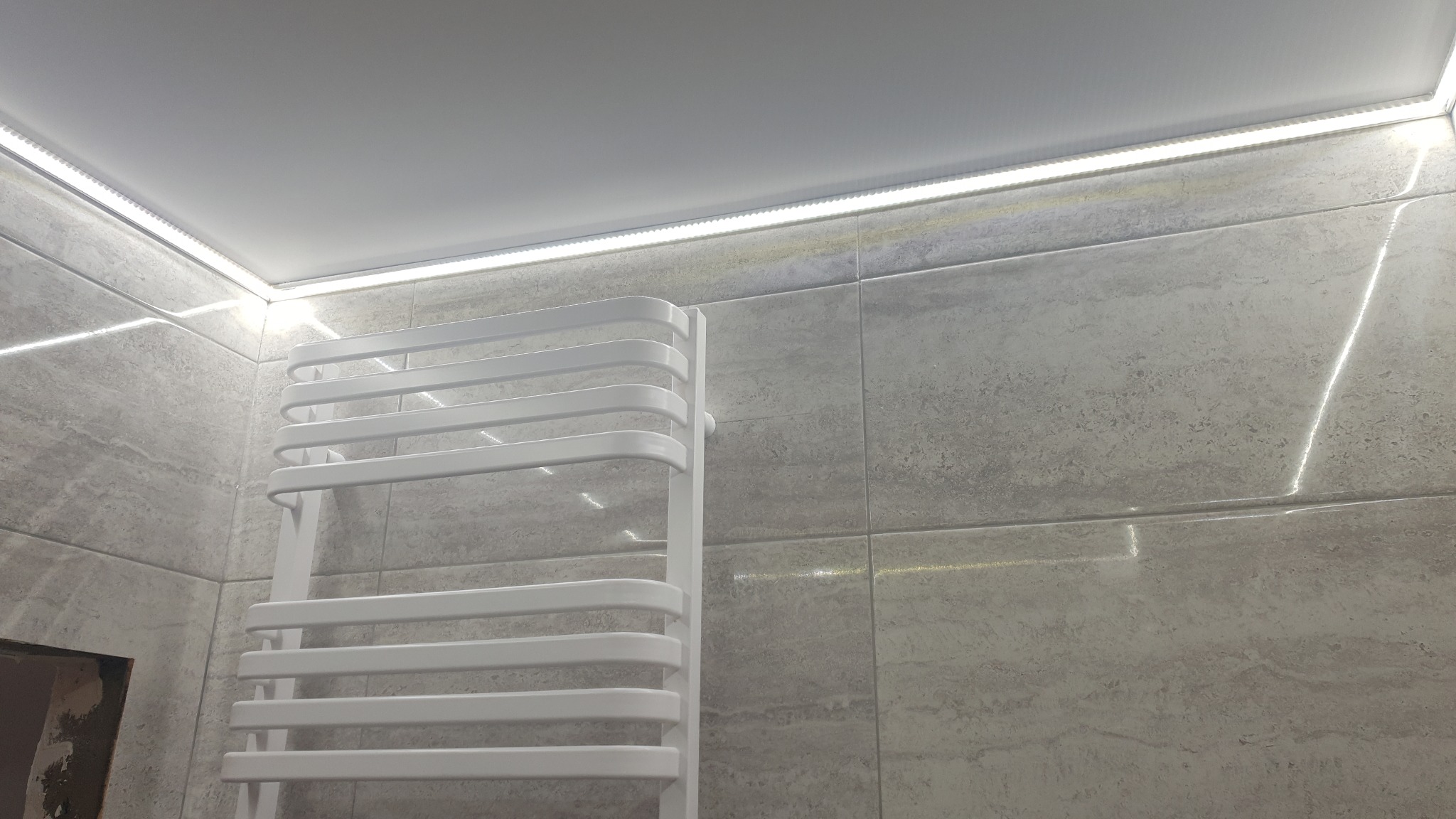 Nowoczesna łazienka z szarymi płytkami imitującymi kamień, podświetlonym sufitem i białym grzejnikiem drabinkowym.
