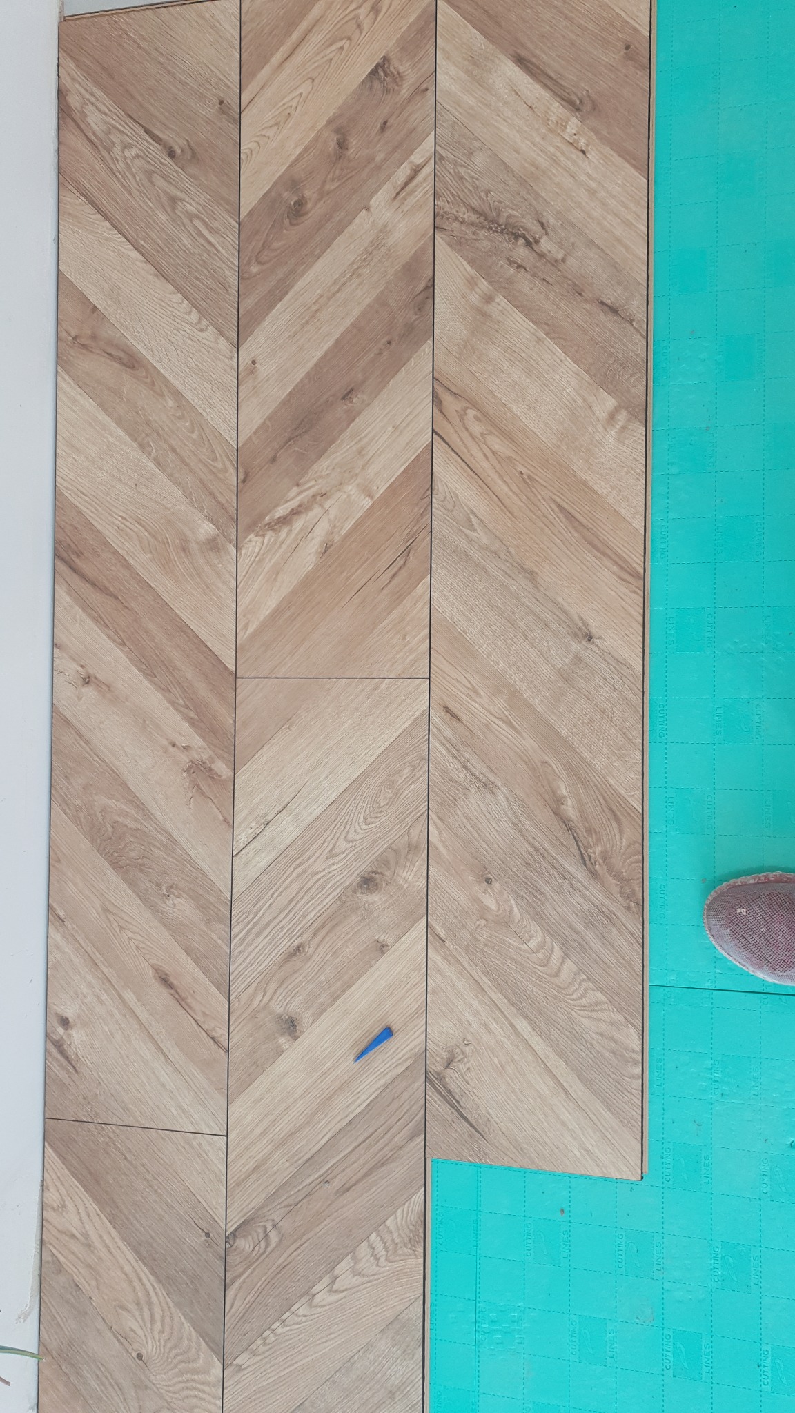 Układanie paneli w jodełkę na zielonej piance izolacyjnej, widoczny niebieski klin dystansowy i fragment buta.