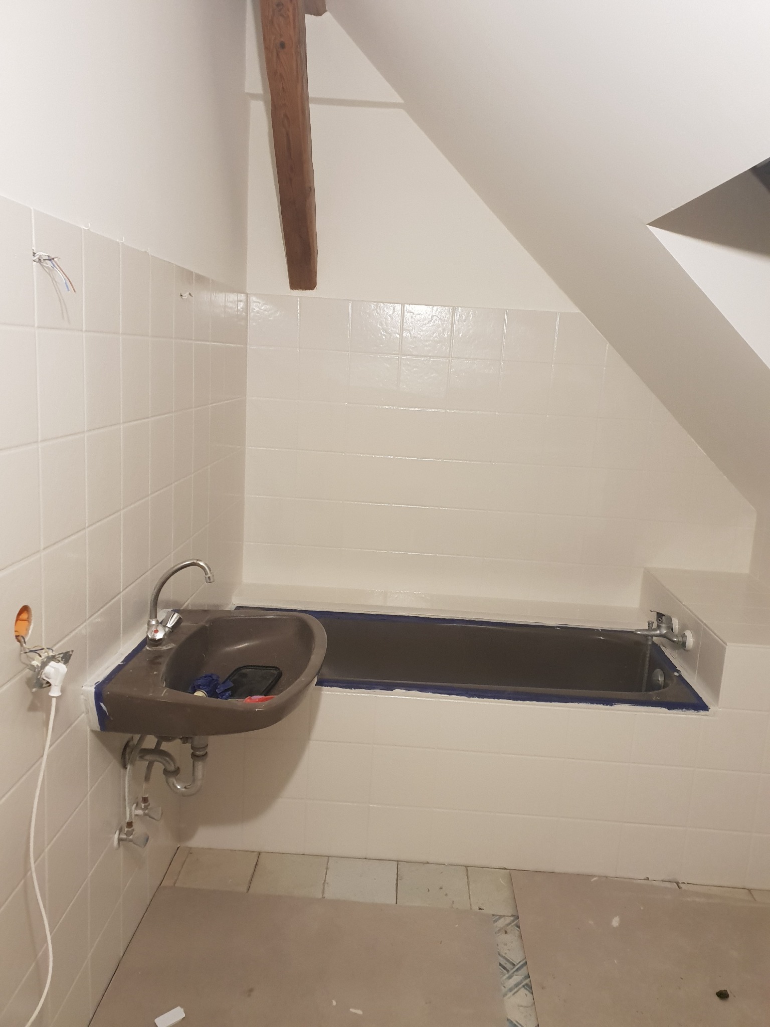 Łazienka w trakcie odświeżania w Warszawie: widoczne zabezpieczone taśmą malarską krawędzie wanny i umywalki, odsłonięte przewody elektryczne i stara glazura na ścianach oraz fragment drewnianej...