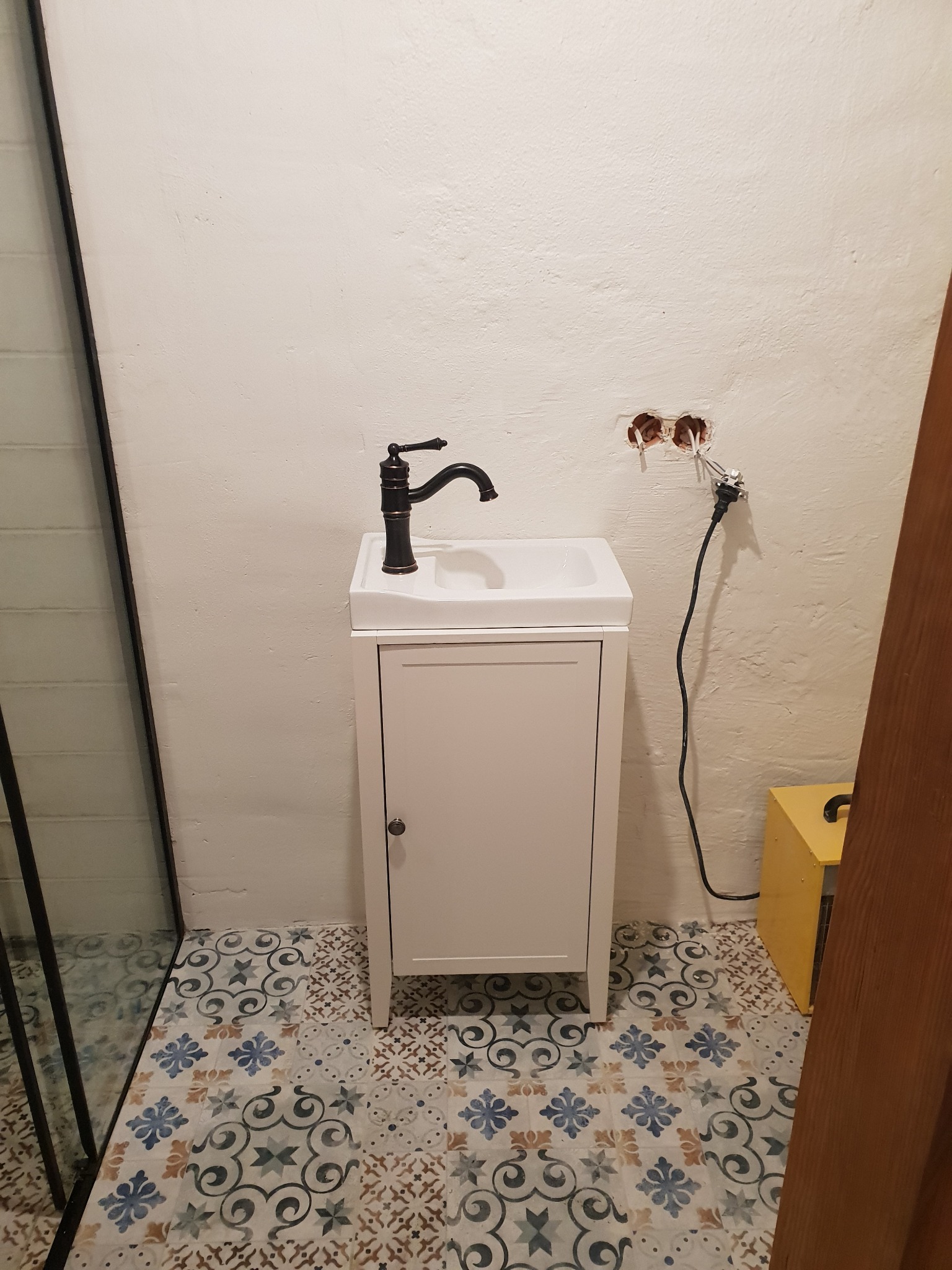 Mała biała szafka pod umywalkę z czarną baterią na tle białej ściany z wystającymi kablami i wzorzystej podłogi z płytek.