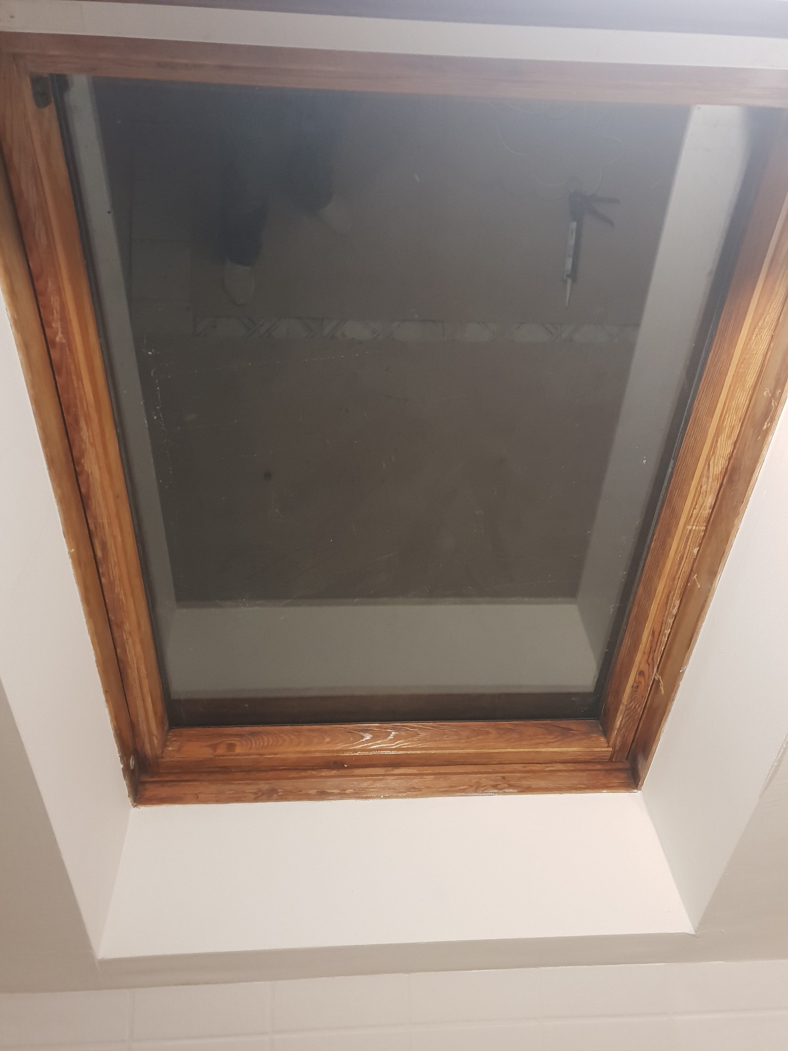 Drewniana rama okna dachowego, widok od dołu, z widocznym aplikatorem silikonu na szybie i odbiciem osoby w górnej części szyby.