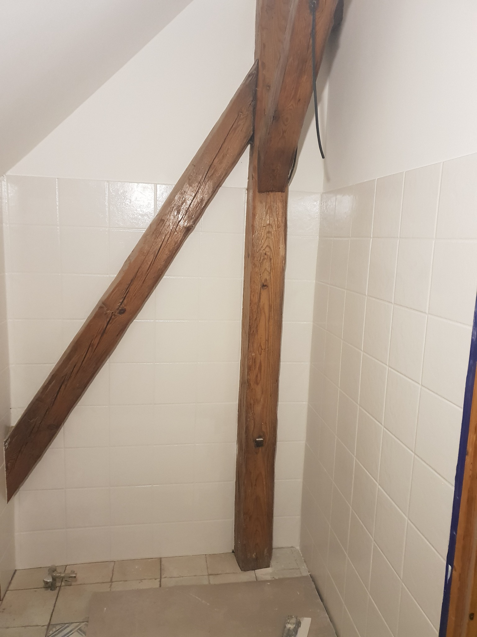 Wnętrze łazienki w trakcie prac: drewniana belka konstrukcyjna na tle białych, kwadratowych płytek ściennych i skośnego sufitu, widoczne narzędzia i fragmenty podłogi w trakcie remontu.