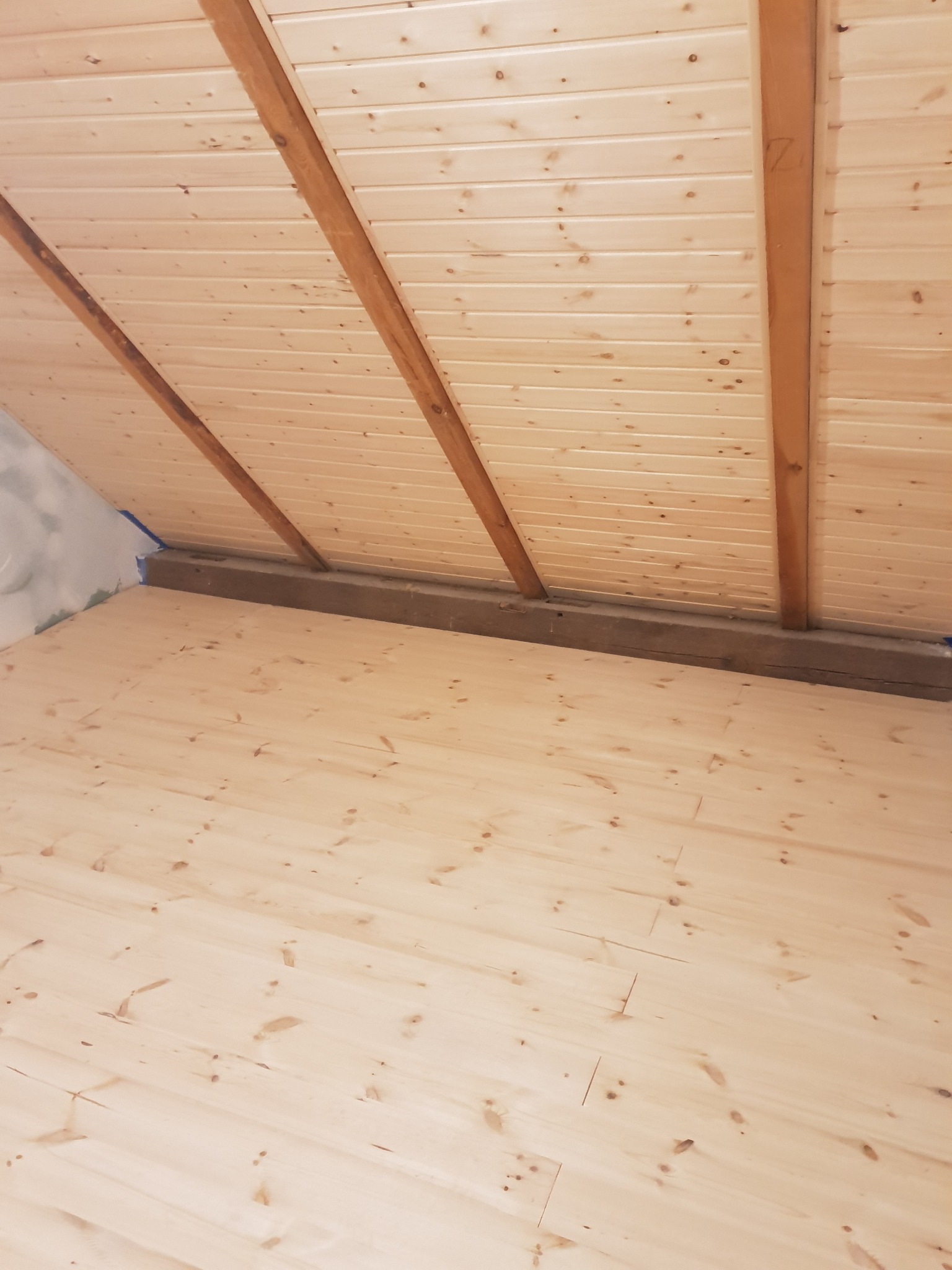 Poddasze z boazerią na skosach dachu i drewnianą podłogą, widoczne ślady taśmy malarskiej na ścianie.