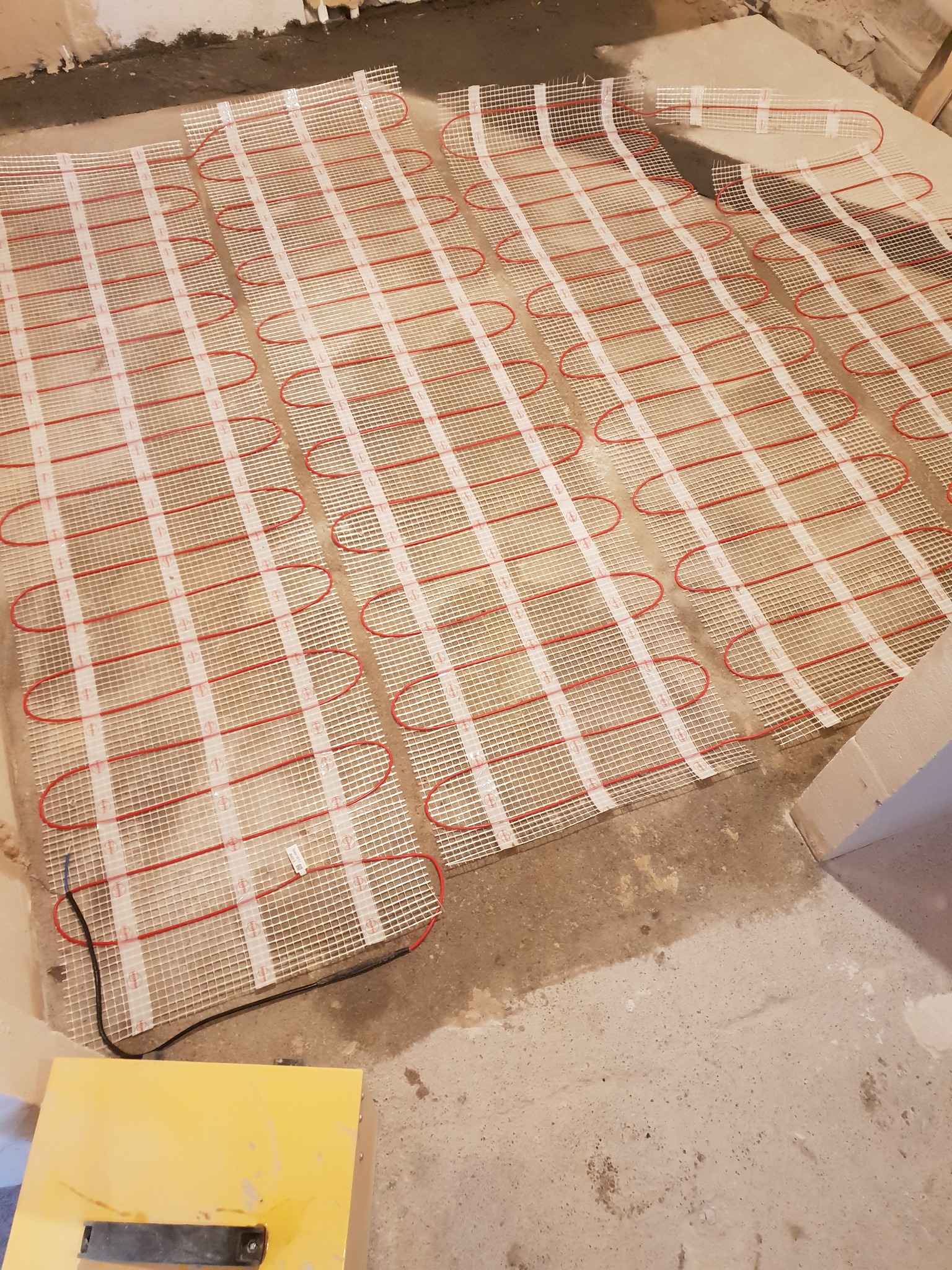 Rozłożona mata grzewcza na podłodze w surowym stanie, widoczne czerwone przewody w siatce, przygotowanie do wylewki.