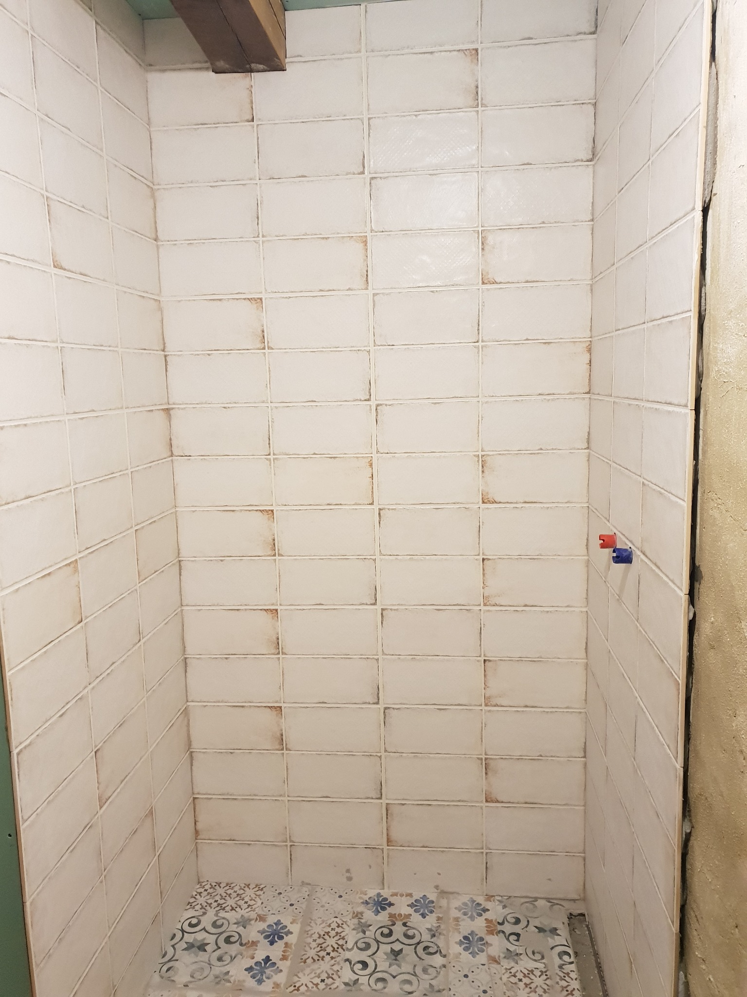 Wnętrze kabiny prysznicowej wyłożonej białymi, prostokątnymi płytkami z delikatnym brązowym obramowaniem, widoczne elementy instalacji wodnej z czerwonym i niebieskim znacznikiem, podłoga...