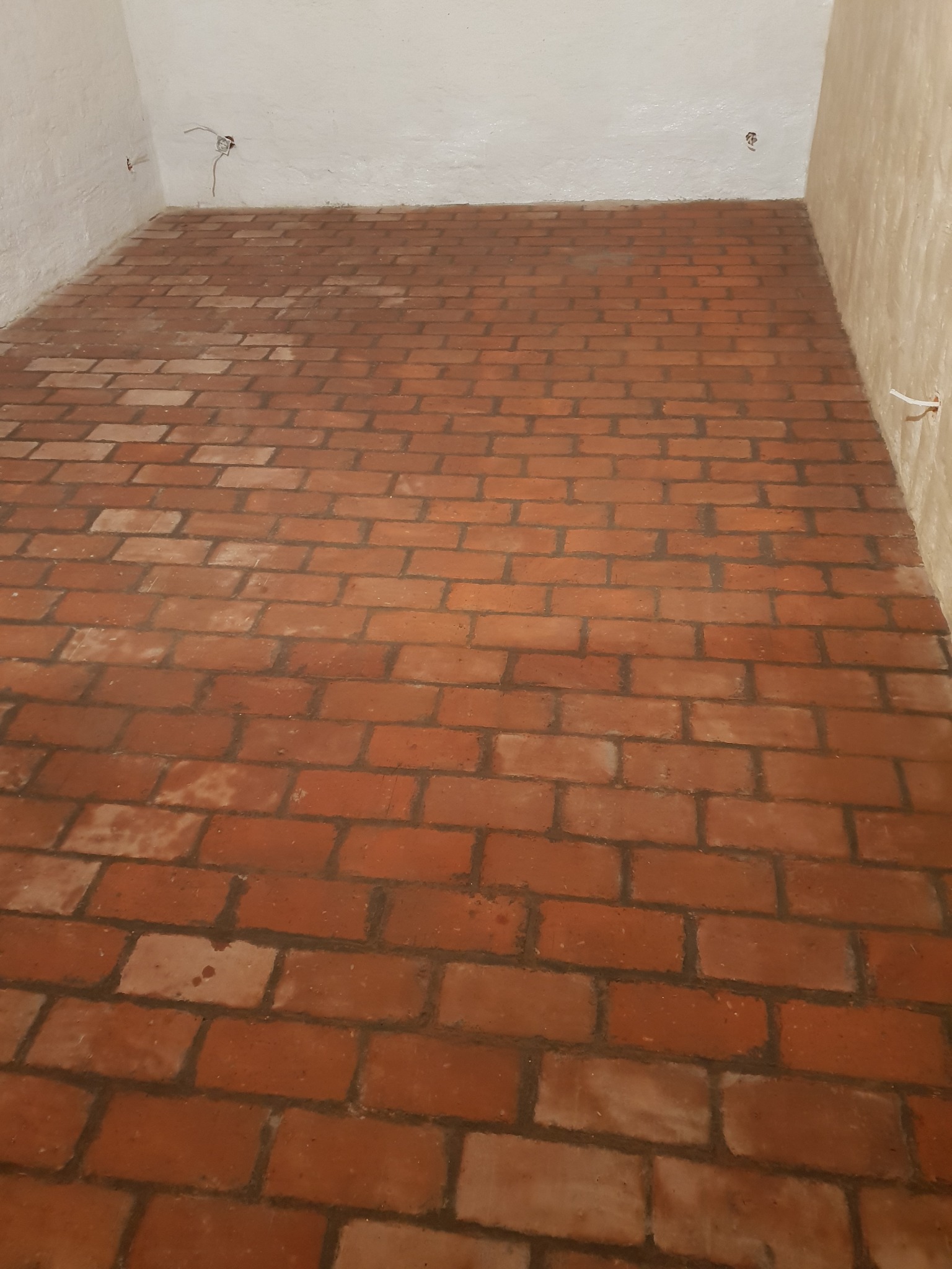 Podłoga wyłożona czerwoną cegłą w pokoju z surowymi, białymi ścianami i wystającymi przewodami elektrycznymi.